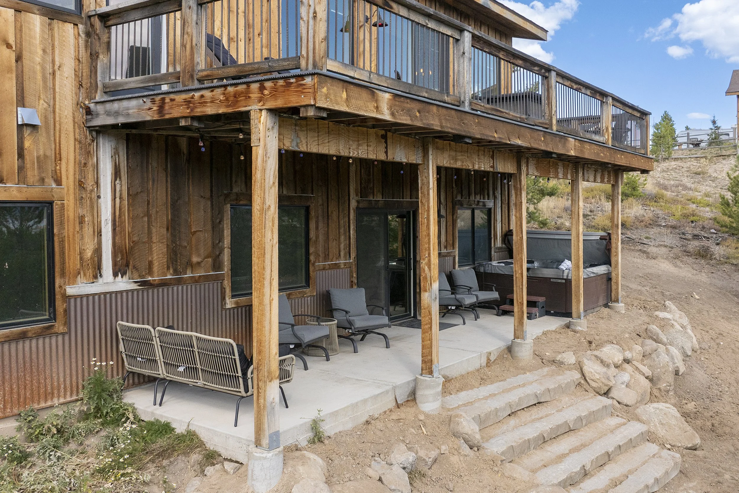 Back porch of Rocky Heart Hideaway chalet in Grand Lake, CO, featuring outdoor chairs, hot tub, and steps leading to the ground, with hillside and trees in the background.