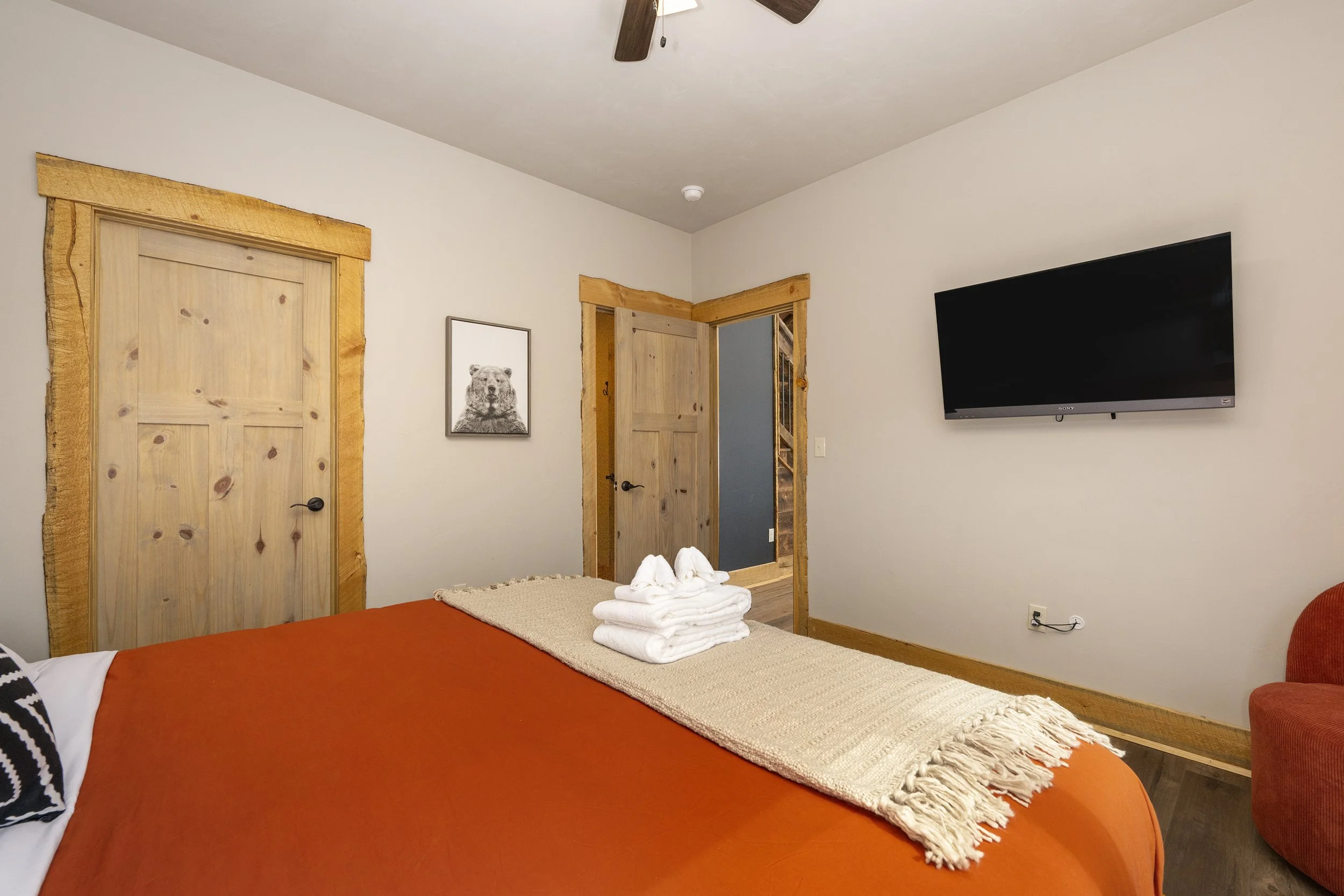 Basement bedroom with a bed covered in an orange blanket, a folded beige towel on the bed, and a landscape scene visible outside through an open door. The room has wooden trim around the doors and a mounted flat-screen TV.