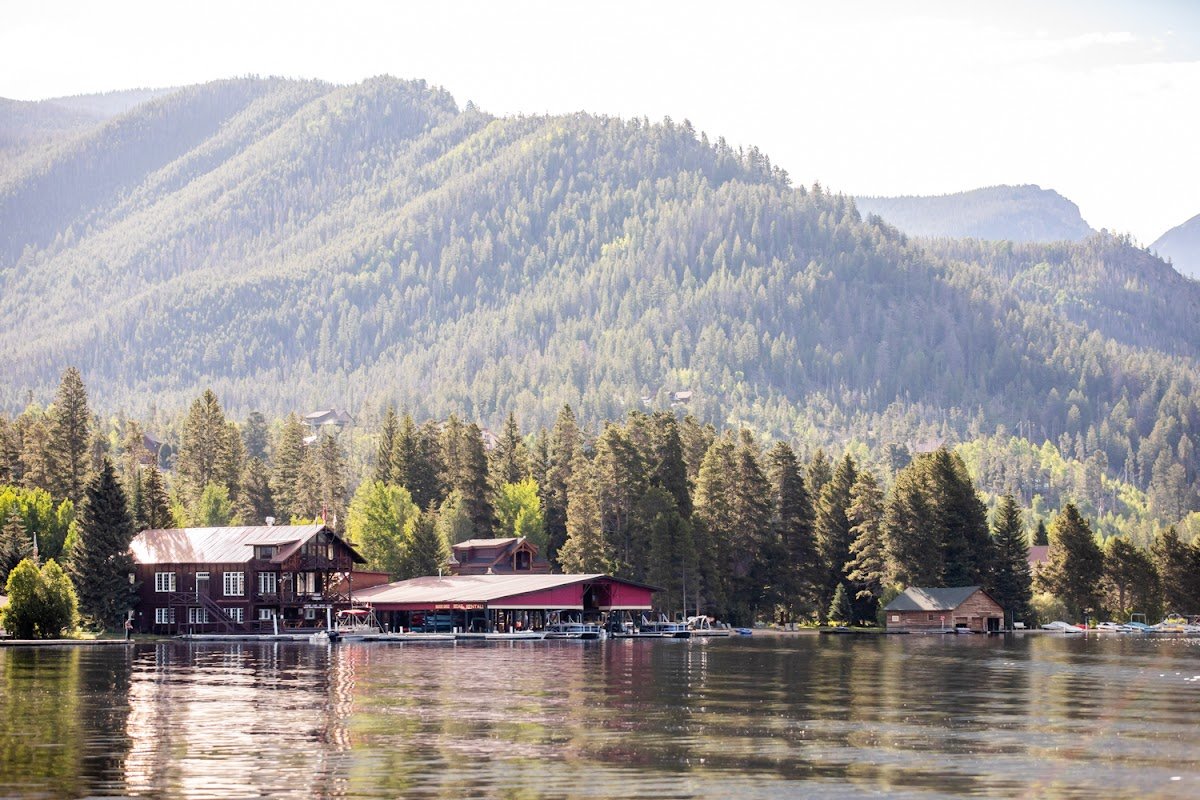 A lakeside scene with a large wooden house and boat docks in the foreground, dense forest and mountains in the background, and calm water reflecting the landscape.