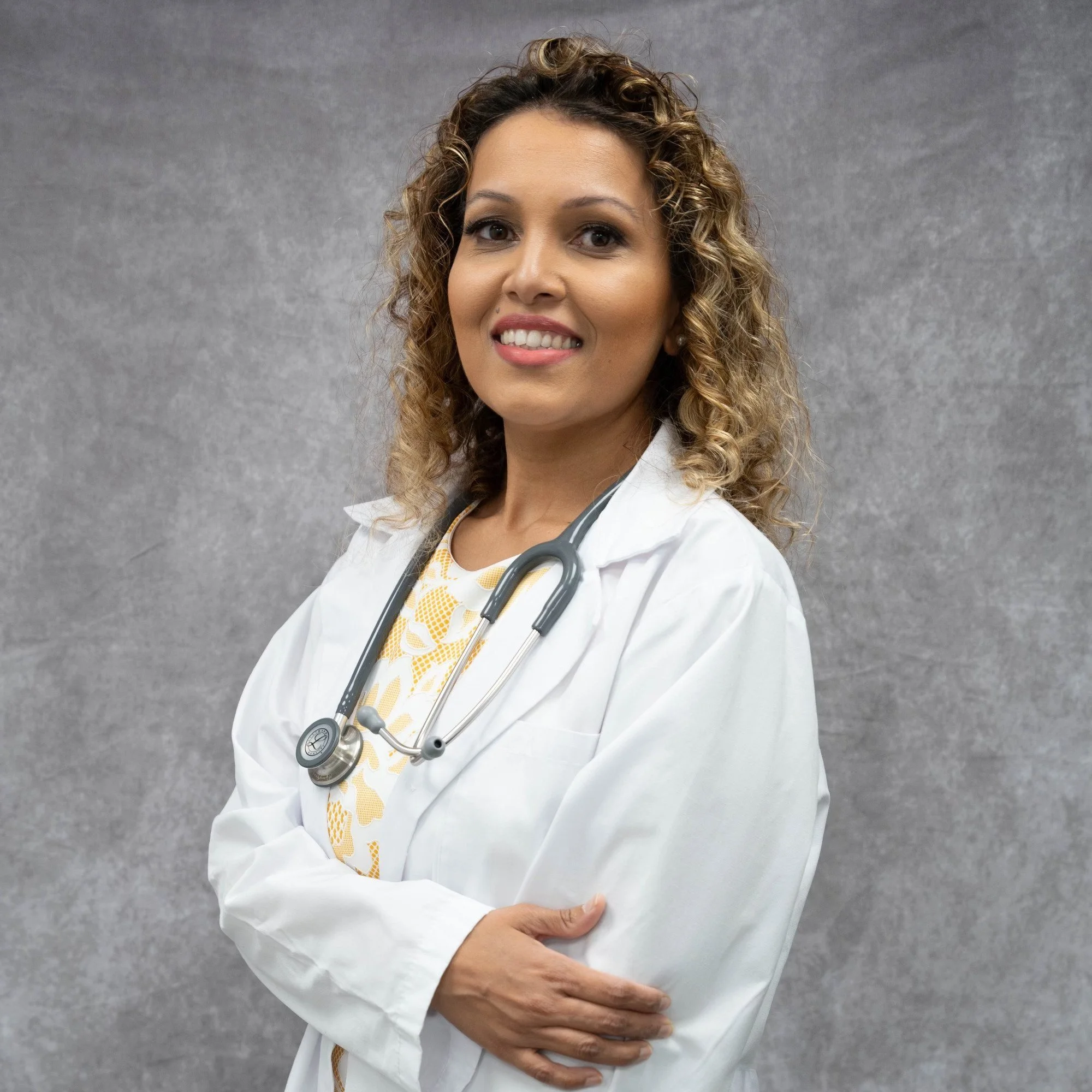 A female doctor with curly hair, wearing a white coat and a stethoscope around her neck, standing against a gray background.