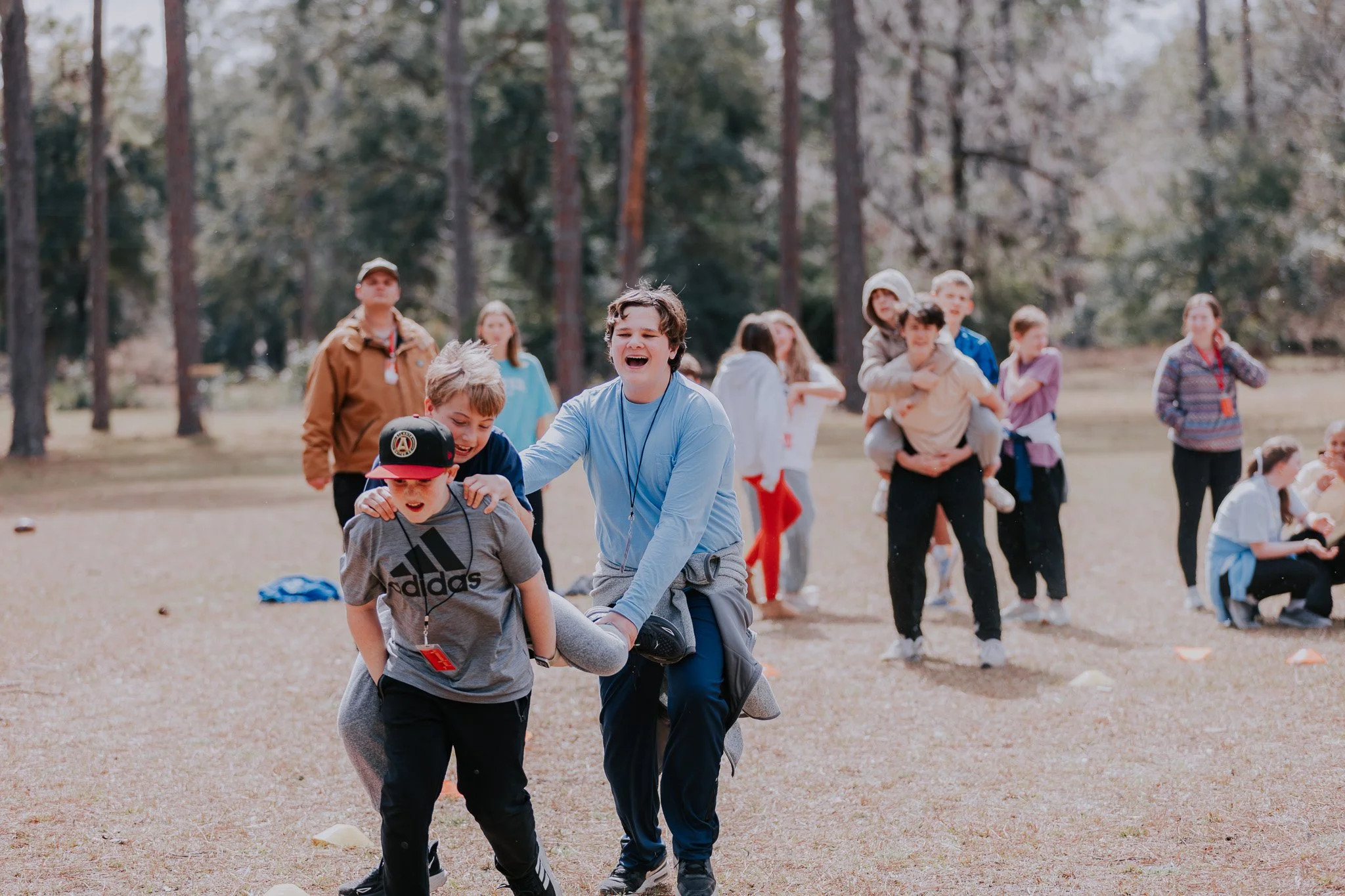 Children and teenagers participating in an outdoor team-building activity in a forested park, with some helping each other cross a line marked on the ground.