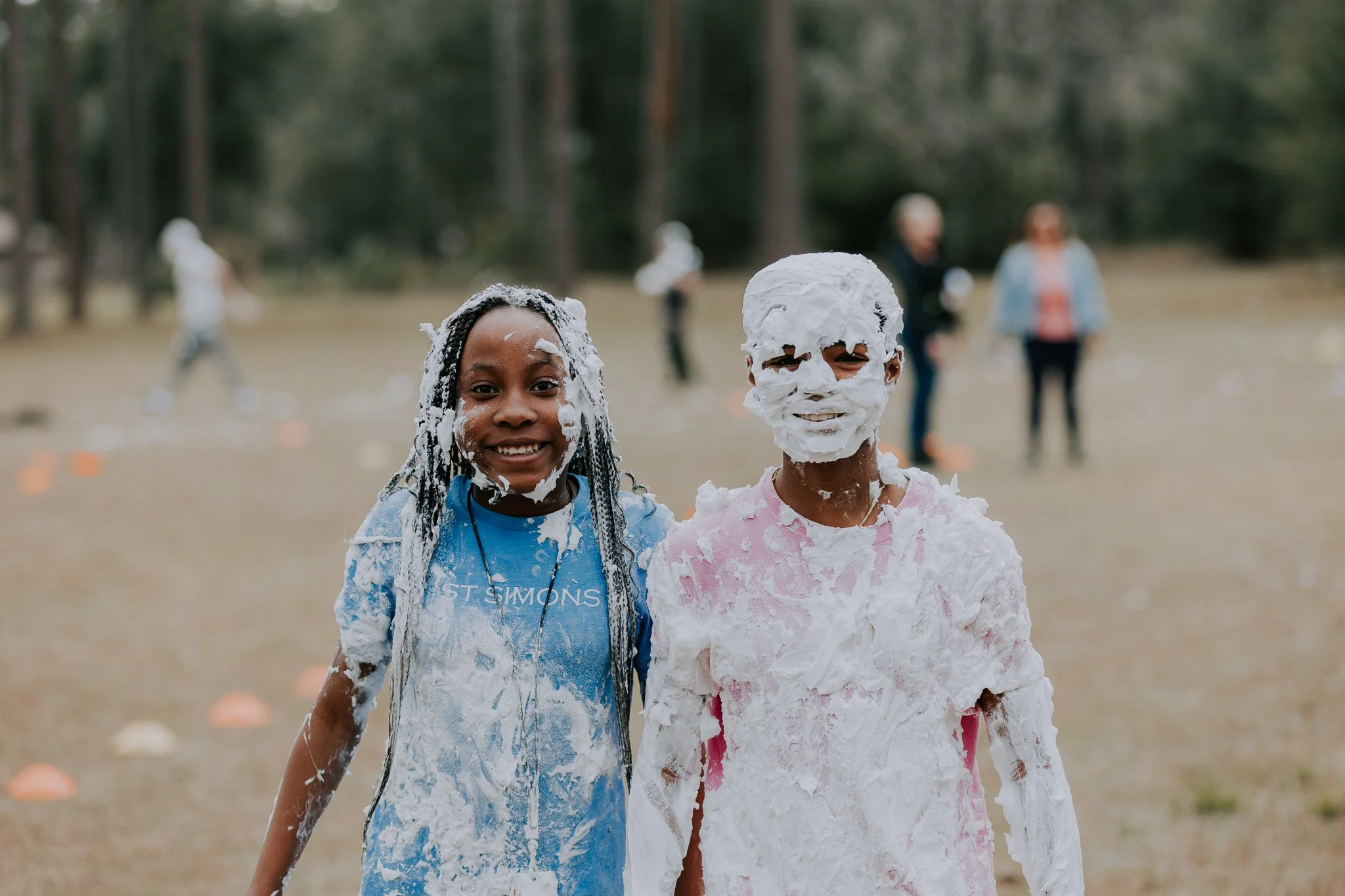 Two children covered in foam smiling outdoors, with other people in the background.