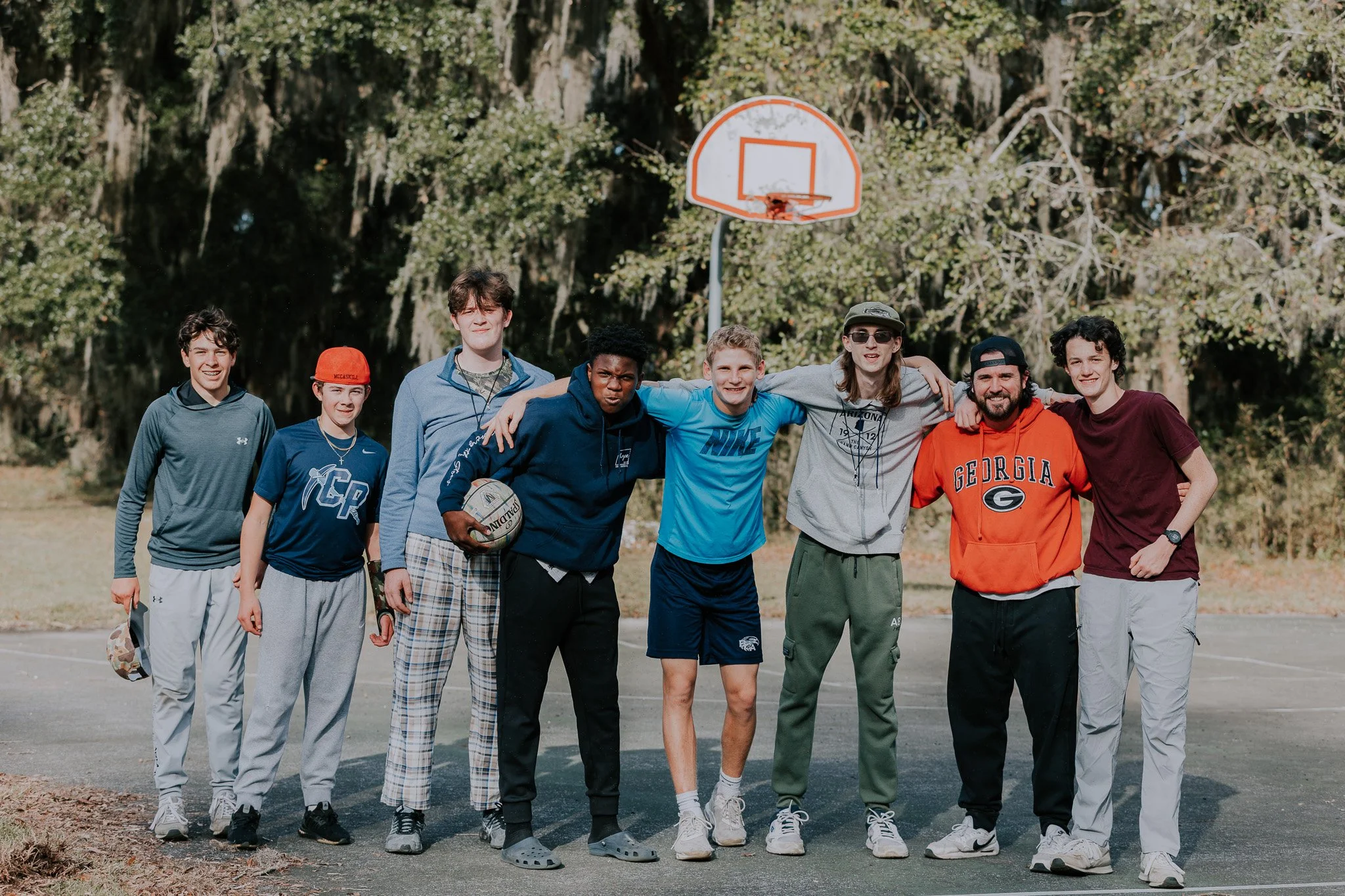 Group of eight boys standing together on an outdoor basketball court, with one holding a basketball, and a basketball hoop in the background.