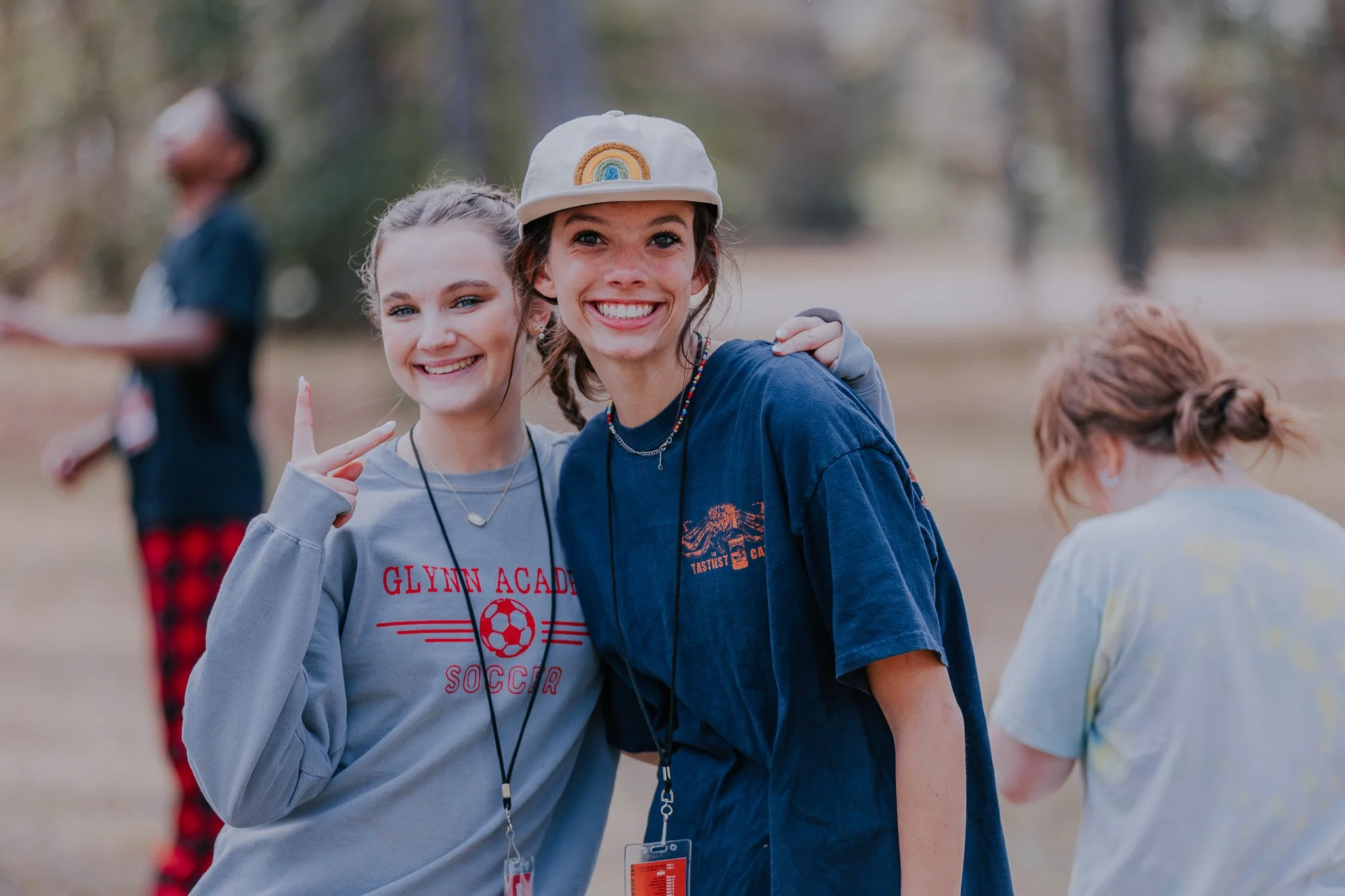 Two smiling girls outdoors, one wearing a grey sweatshirt with 'Glynn Academy Soccer' and the other in a navy T-shirt with a rainbow cap, posing with their arms around each other, with a blurred person in the background.