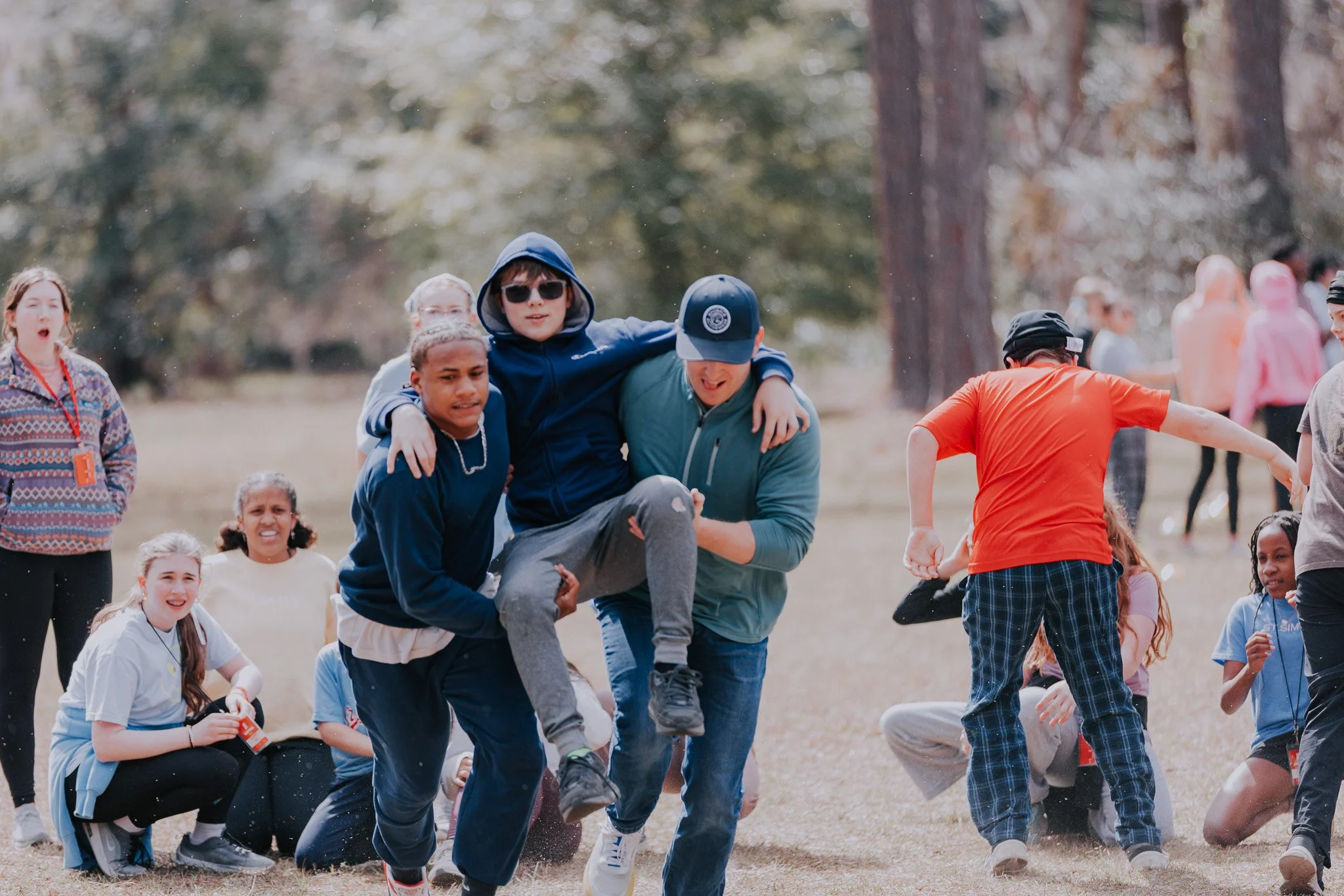 Group of children and teenagers outdoors in a field, engaging in a playful activity, with some sitting on the ground and others standing or leaning, surrounded by trees in the background.