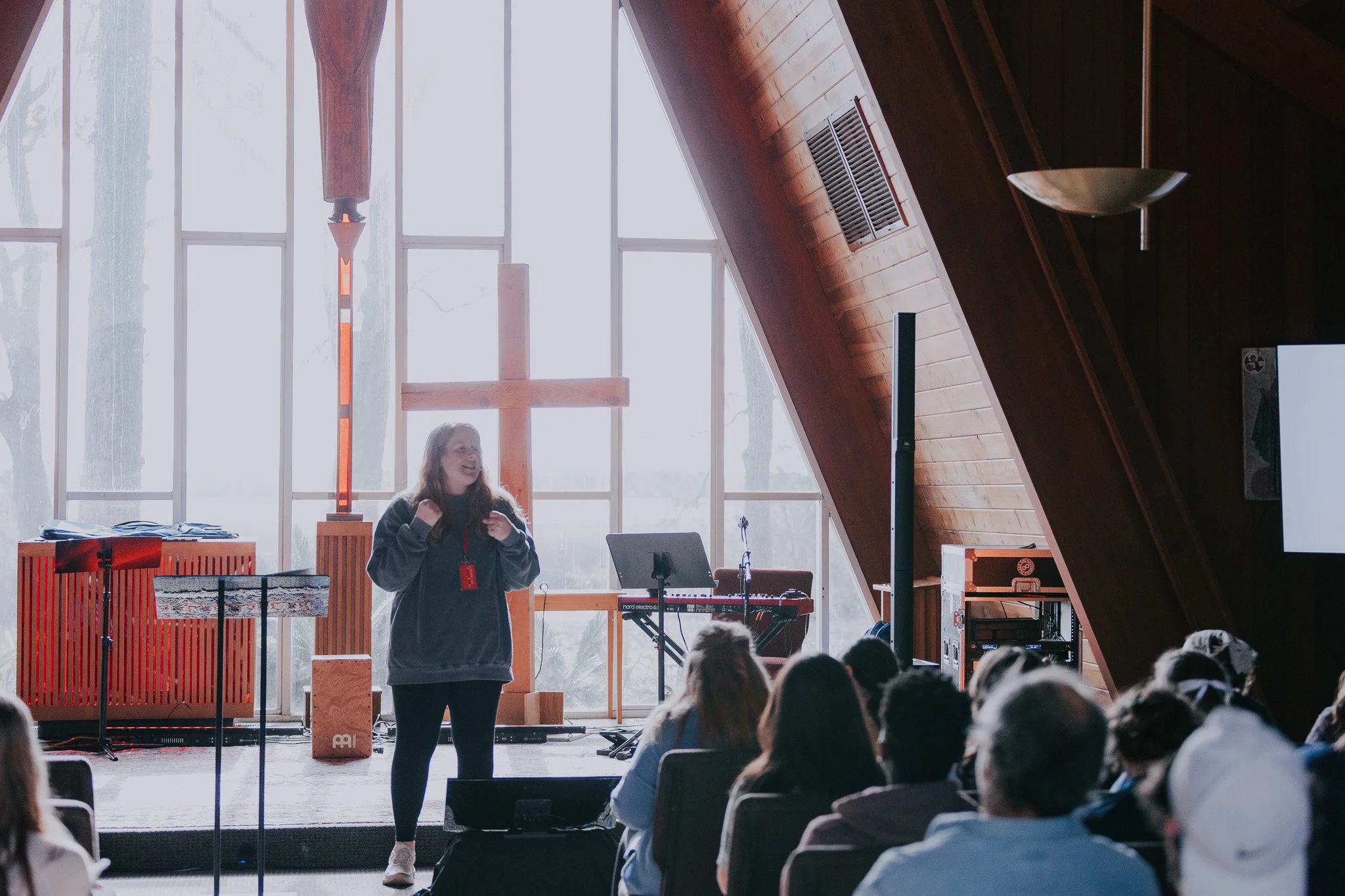 Young woman with a lanyard speaking to an audience in a church or chapel, with a large wooden cross and a stained glass window behind her.