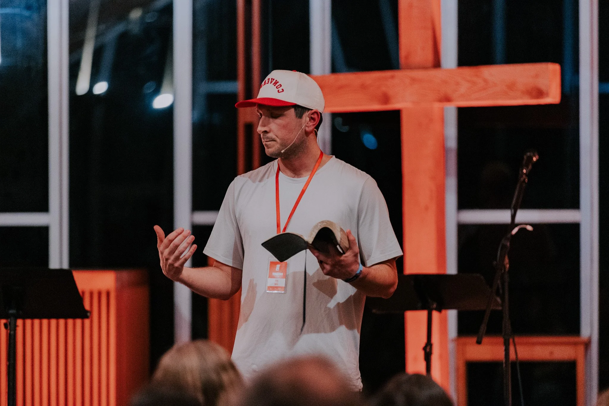 A man speaking at an event, wearing a white t-shirt, a white and red cap, and holding a small book. There are microphones and orange framed structures in the background.