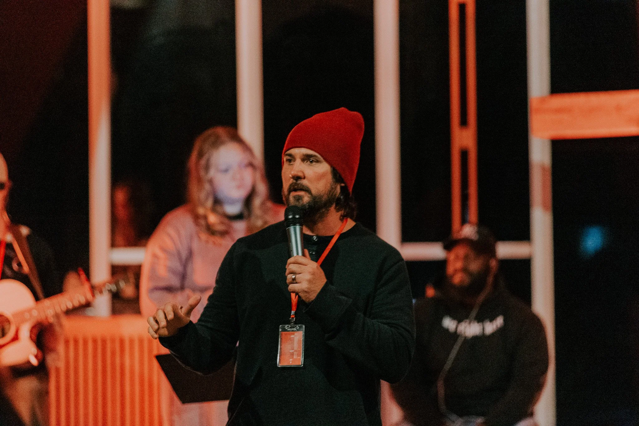 A man with long dark hair and a beard, wearing a red beanie and black shirt, speaks into a microphone on stage during a performance or event, with a woman with long blonde hair and light-colored top in the background, and a person playing guitar, all