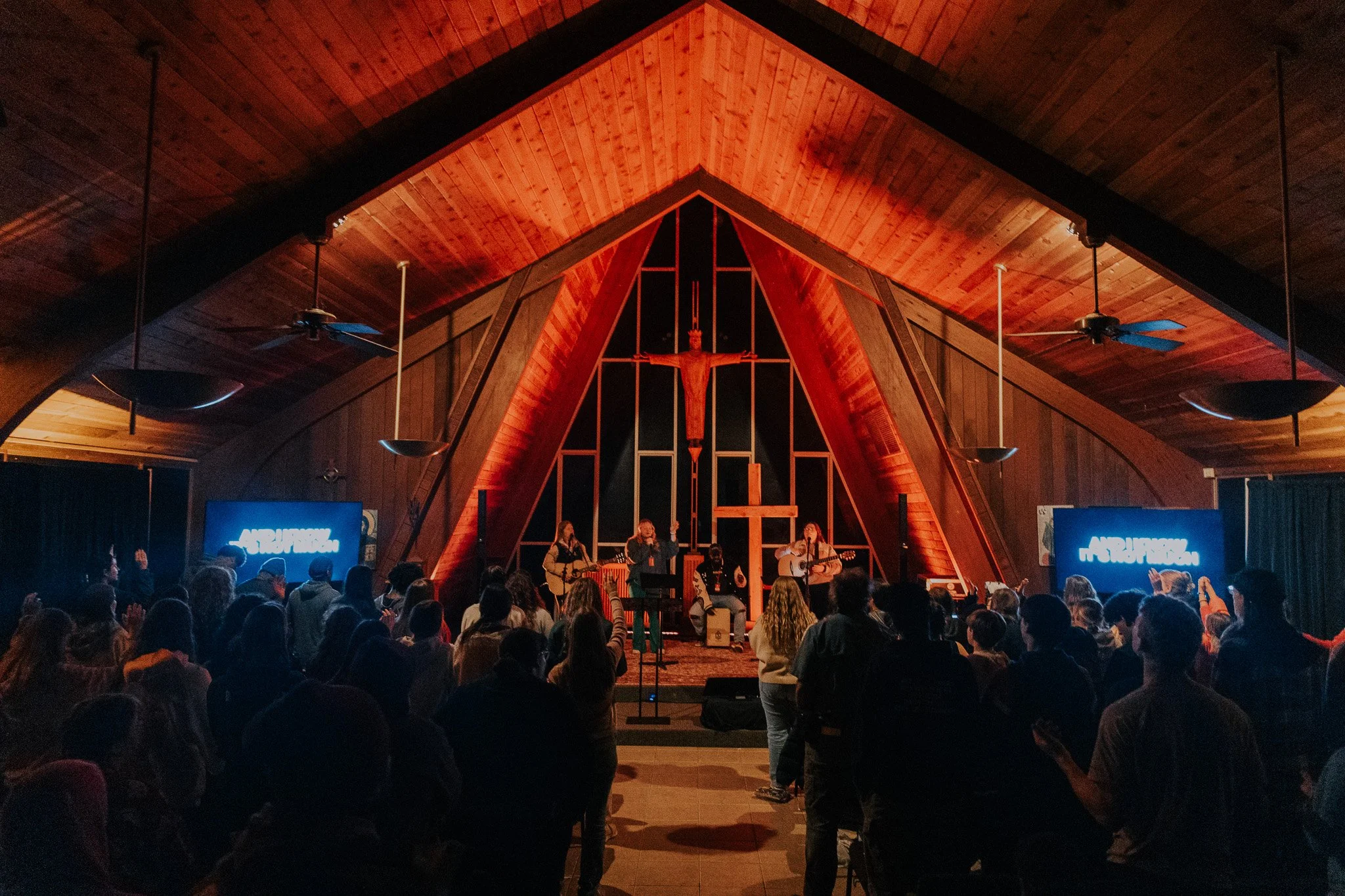 A church interior with wooden ceiling and walls, featuring a cross with a crucifix and a crucified Jesus at the altar. A band of musicians is performing on stage. The congregation is standing, some clapping or raising hands, and there are two screens