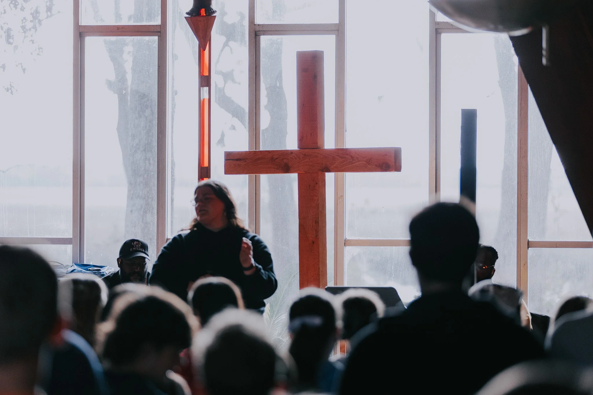 People gathered in a church service with a large wooden cross at the front, backlit by natural light from large windows.