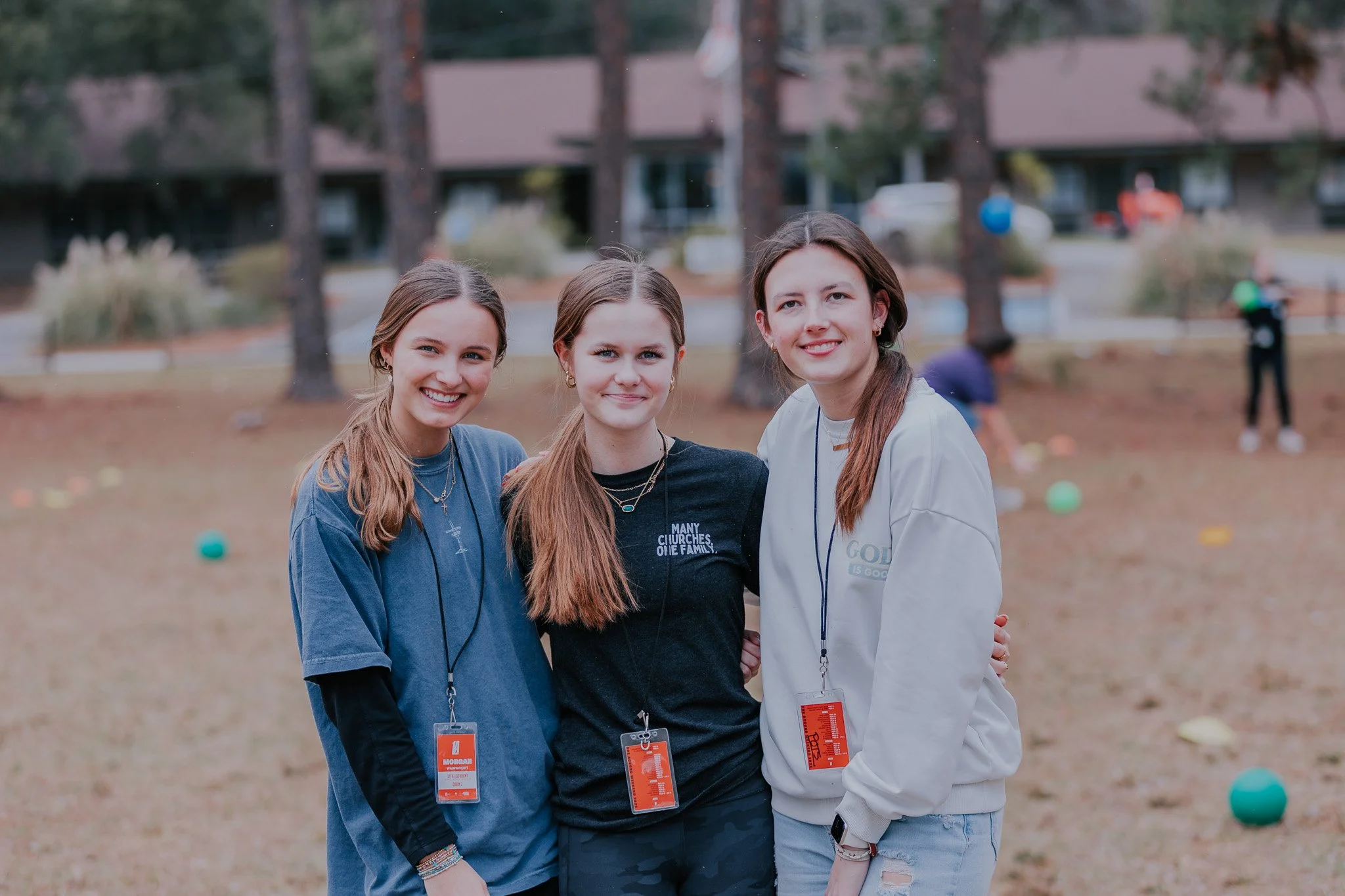 Three young women standing together outdoors in a park, smiling at the camera. They are wearing casual clothing and have name tags around their necks. The background shows trees, people, and colorful balls on the ground.