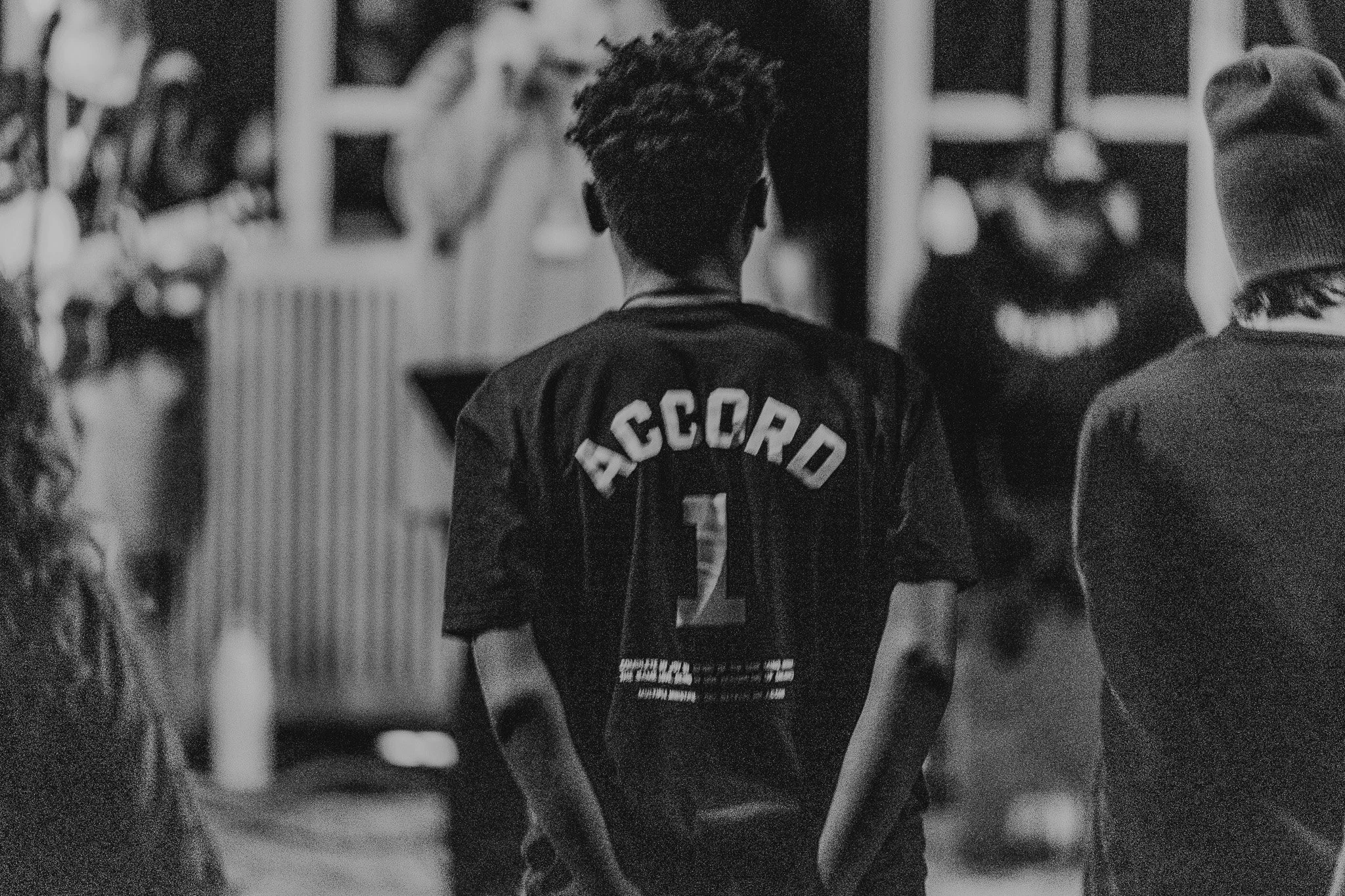 A young person with short, curly hair is walking outdoors, wearing an 'ACCORD' sports jersey with the number 1 on the back, surrounded by several other people.