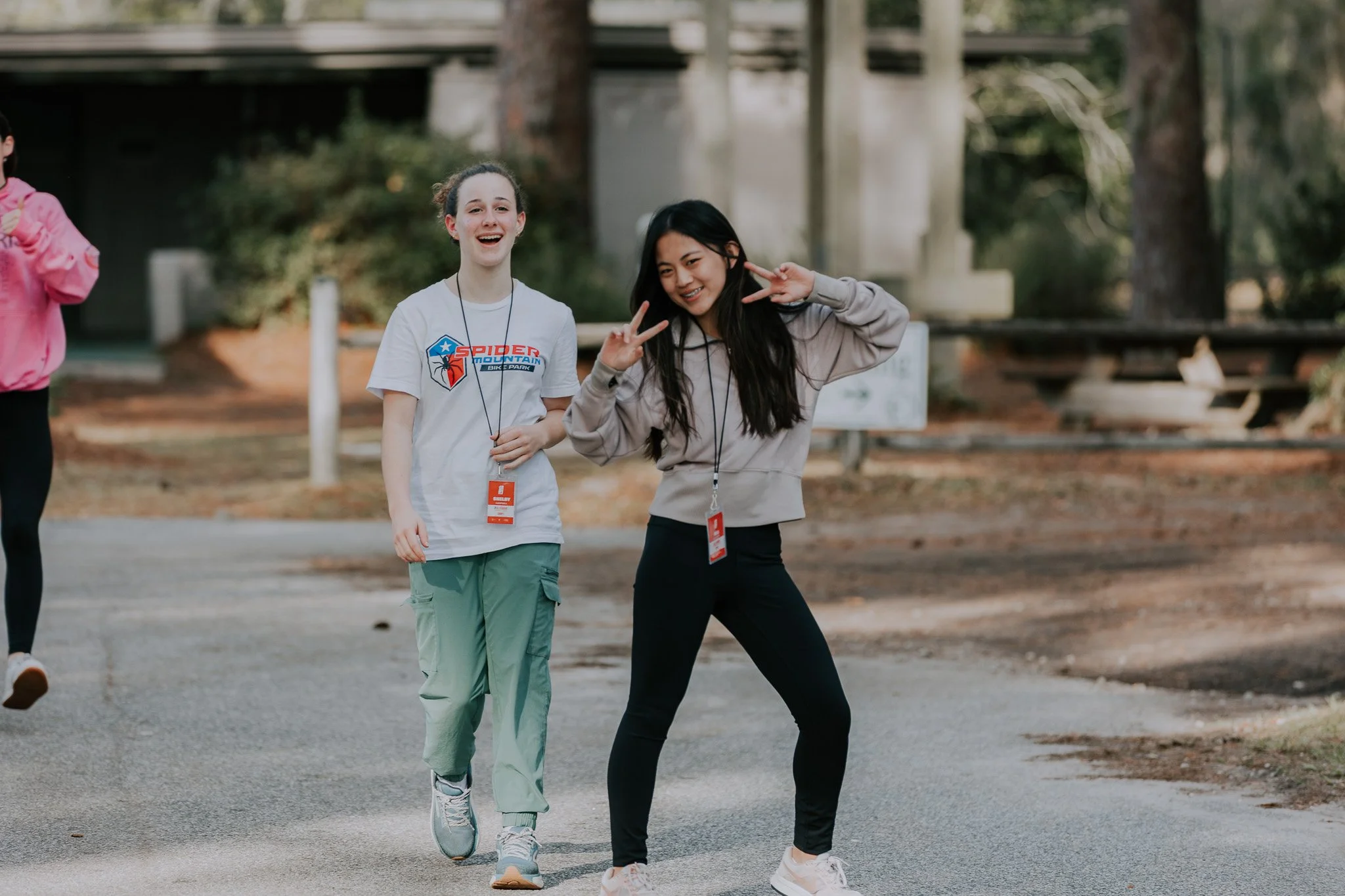 Two young women smiling and posing with peace signs, walking outdoors in a park with trees and wooden picnic tables in the background.