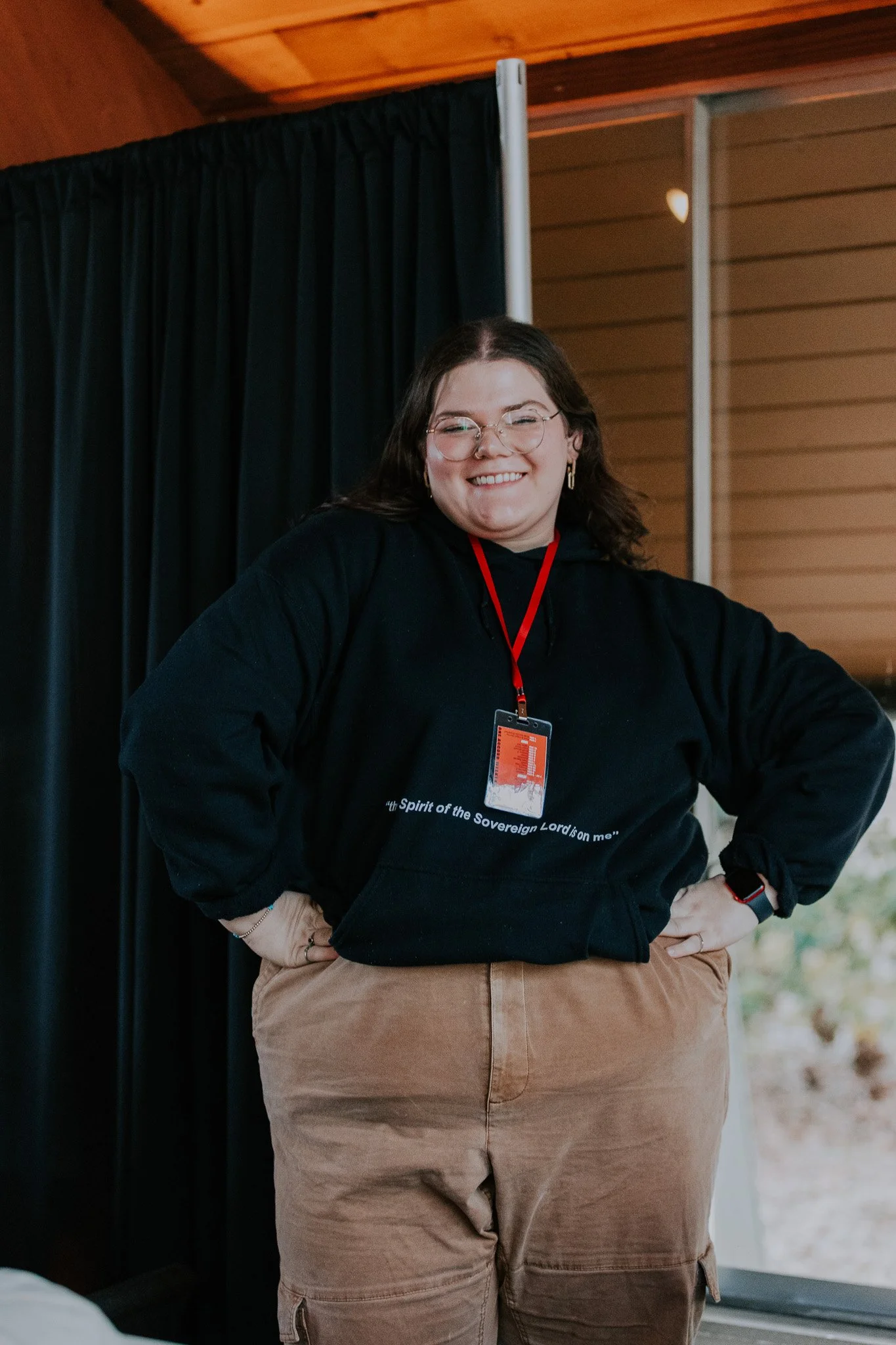 A woman with glasses, wearing a black hoodie with white text, tan cargo pants, and a red lanyard with a pass, smiling with hands on hips in front of black curtains in an indoor setting.