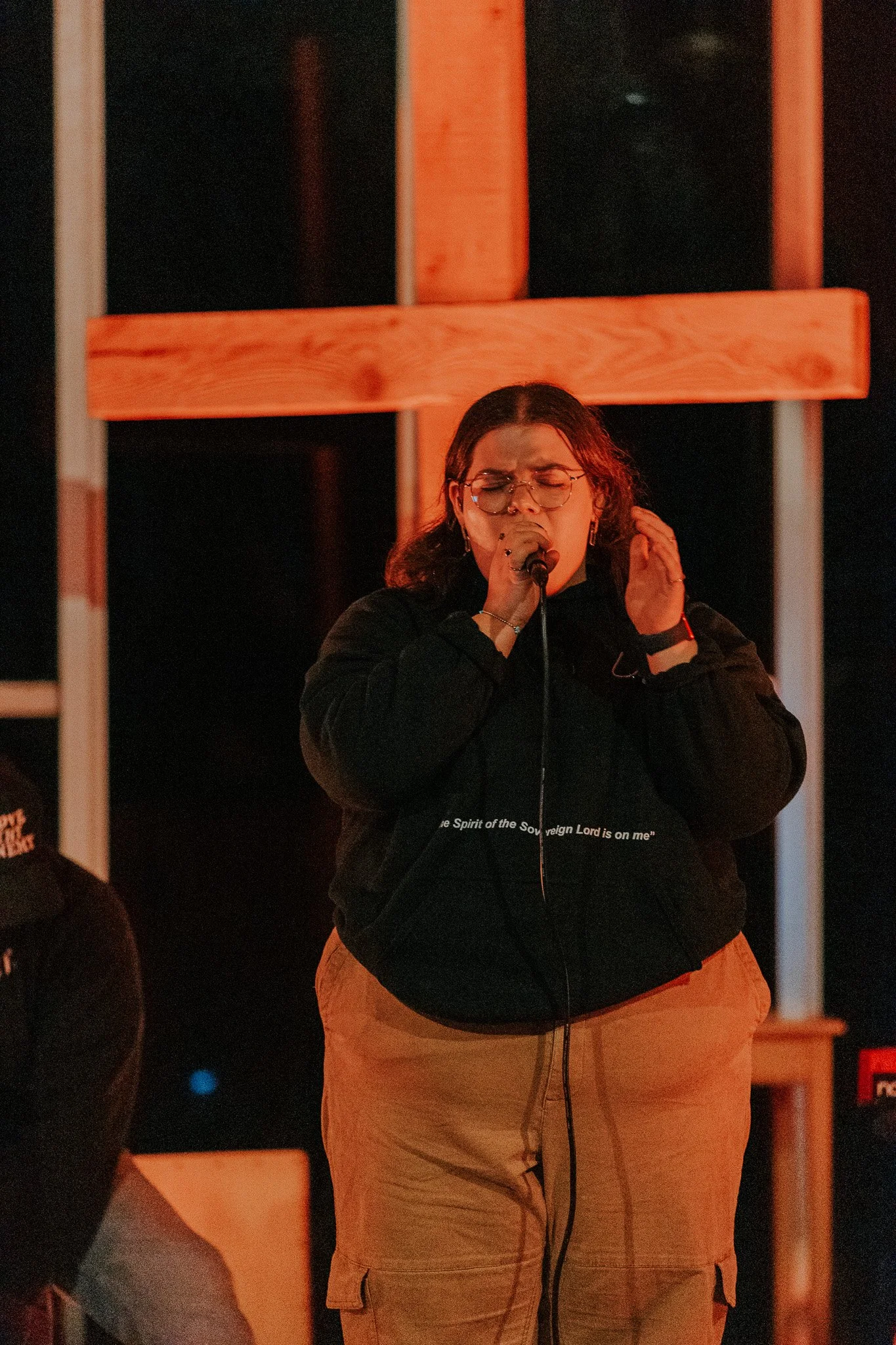 A woman singing into a microphone with her eyes closed, wearing glasses, a black hoodie, and beige cargo pants, standing in front of a wooden cross.