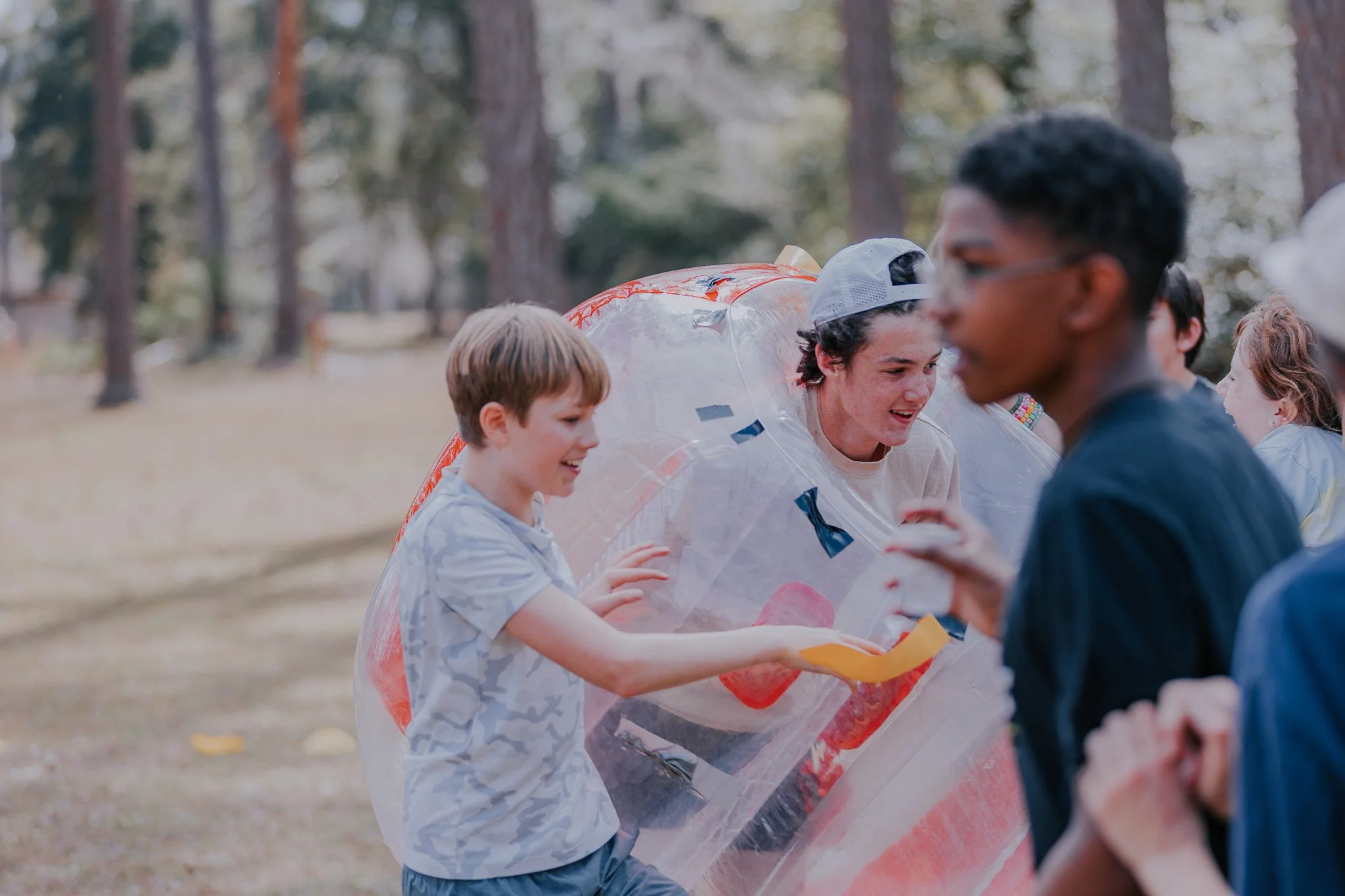 Kids playing with a large inflatable bubble outdoors in a wooded park during daytime.