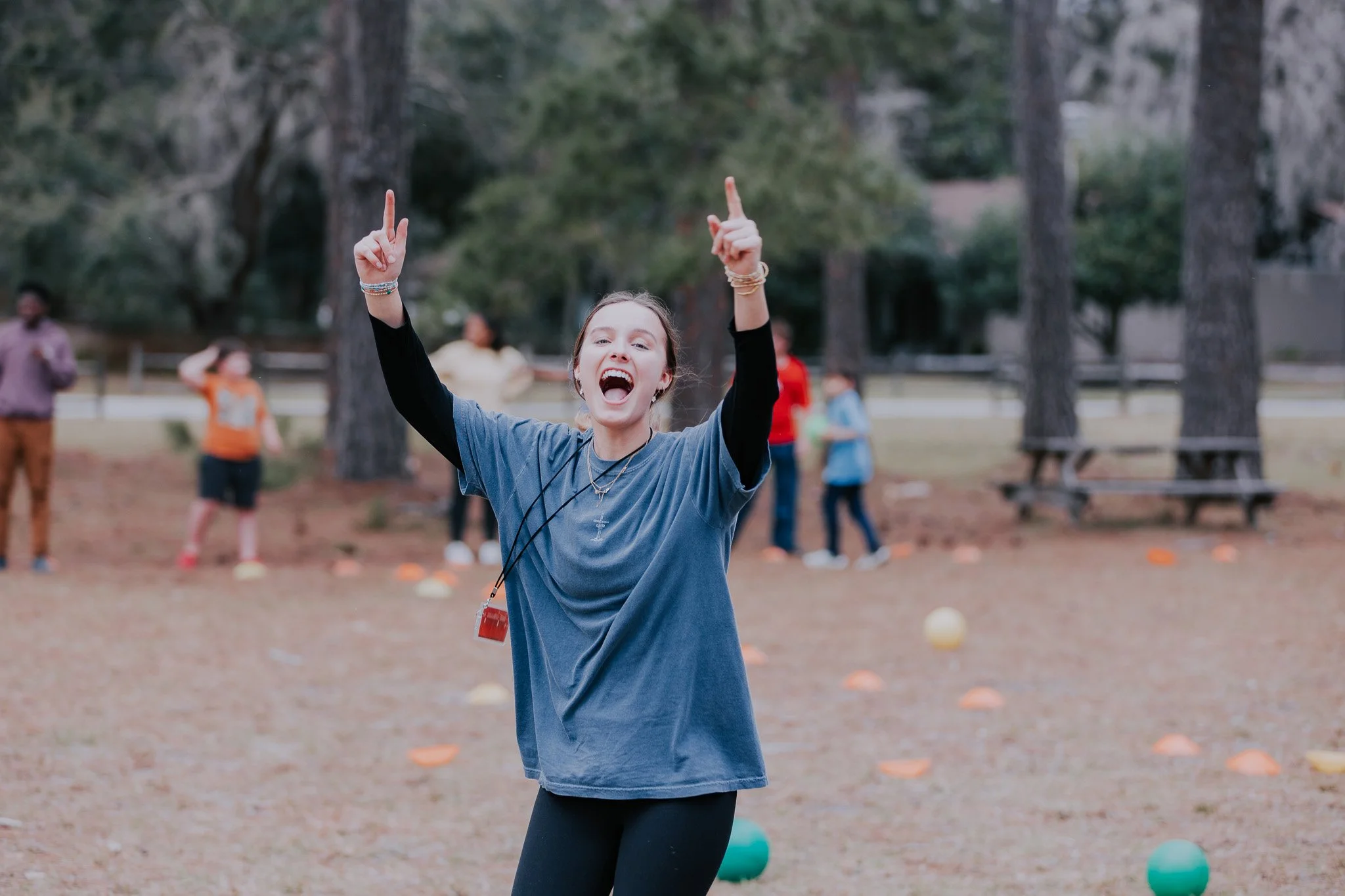 A young woman with arms raised and mouth open in excitement on a field with trees, with several other people in the background.