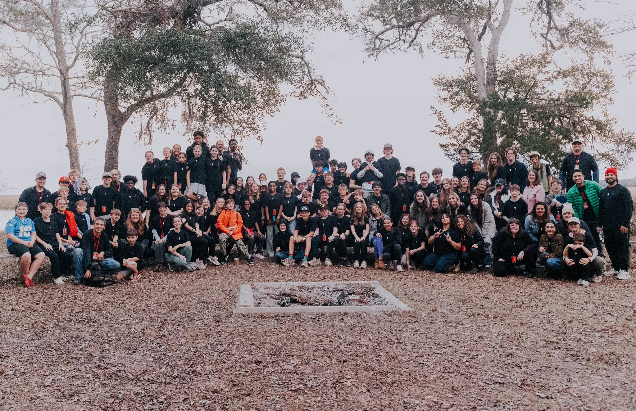 A large group of people, including children and adults, are gathered outdoors for a group photo around a fire pit under tall trees with sparse leaves. Many are wearing black shirts, some have red lanyards, and a few are in different outfits. The scen
