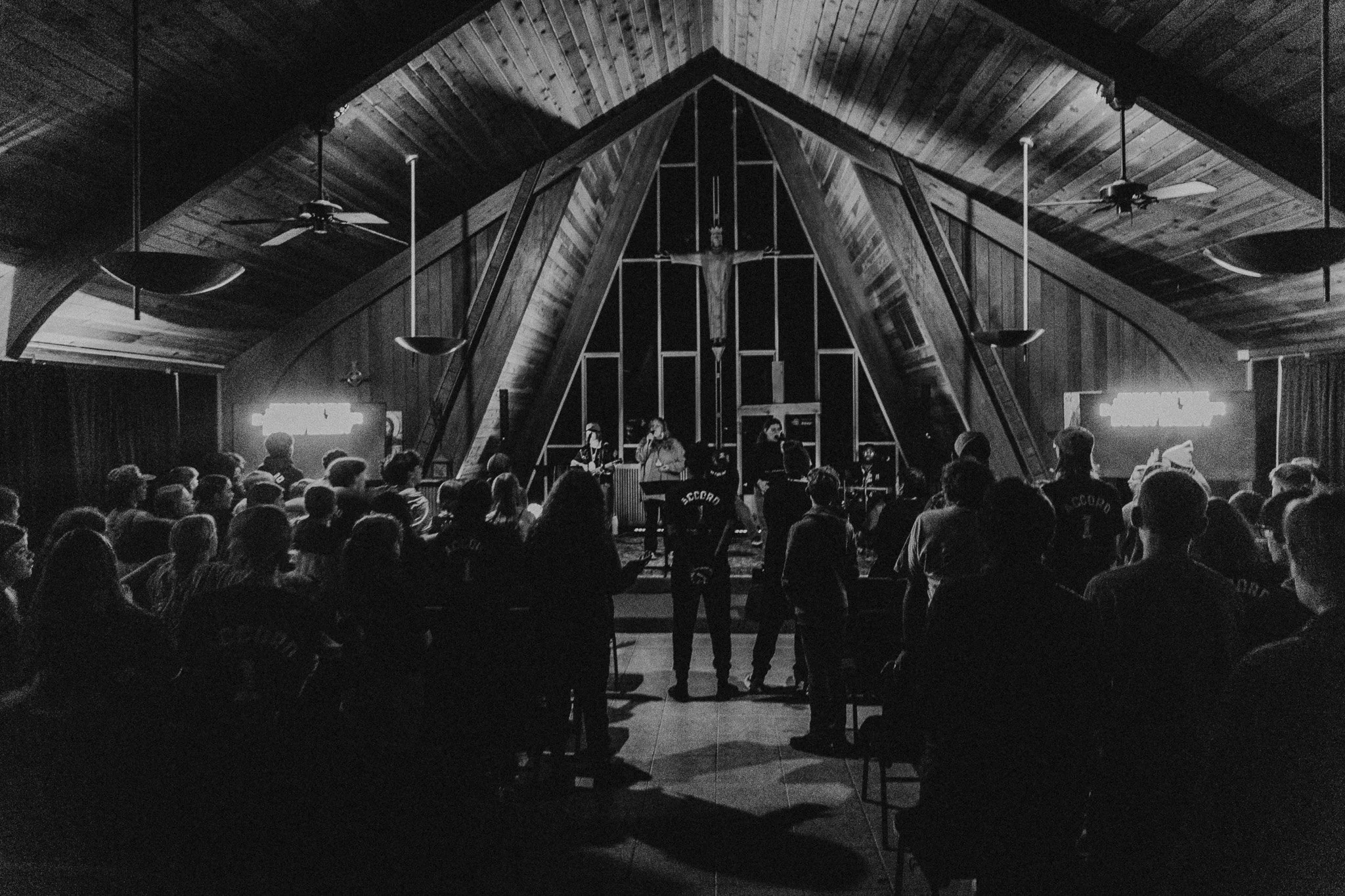 A black and white photo of a church interior with a congregation gathered for a service. The altar has a crucifix and a band playing music. Audience members are standing and seated, some wearing uniforms that read 'RECOR'.