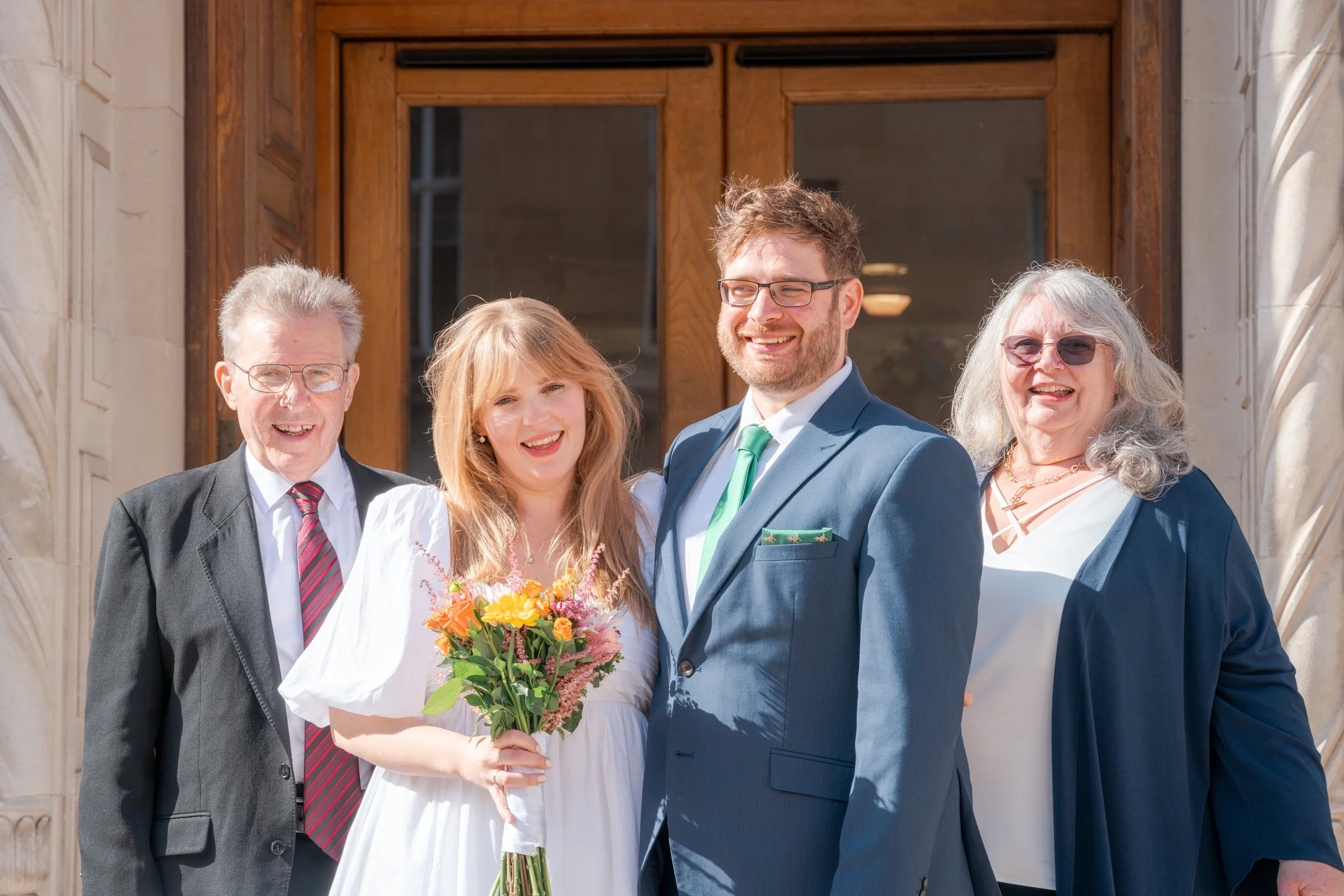 A wedding party standing outside a building with wooden doors, including a bride holding a bouquet of flowers, a groom in a suit, and three older adults, all smiling.