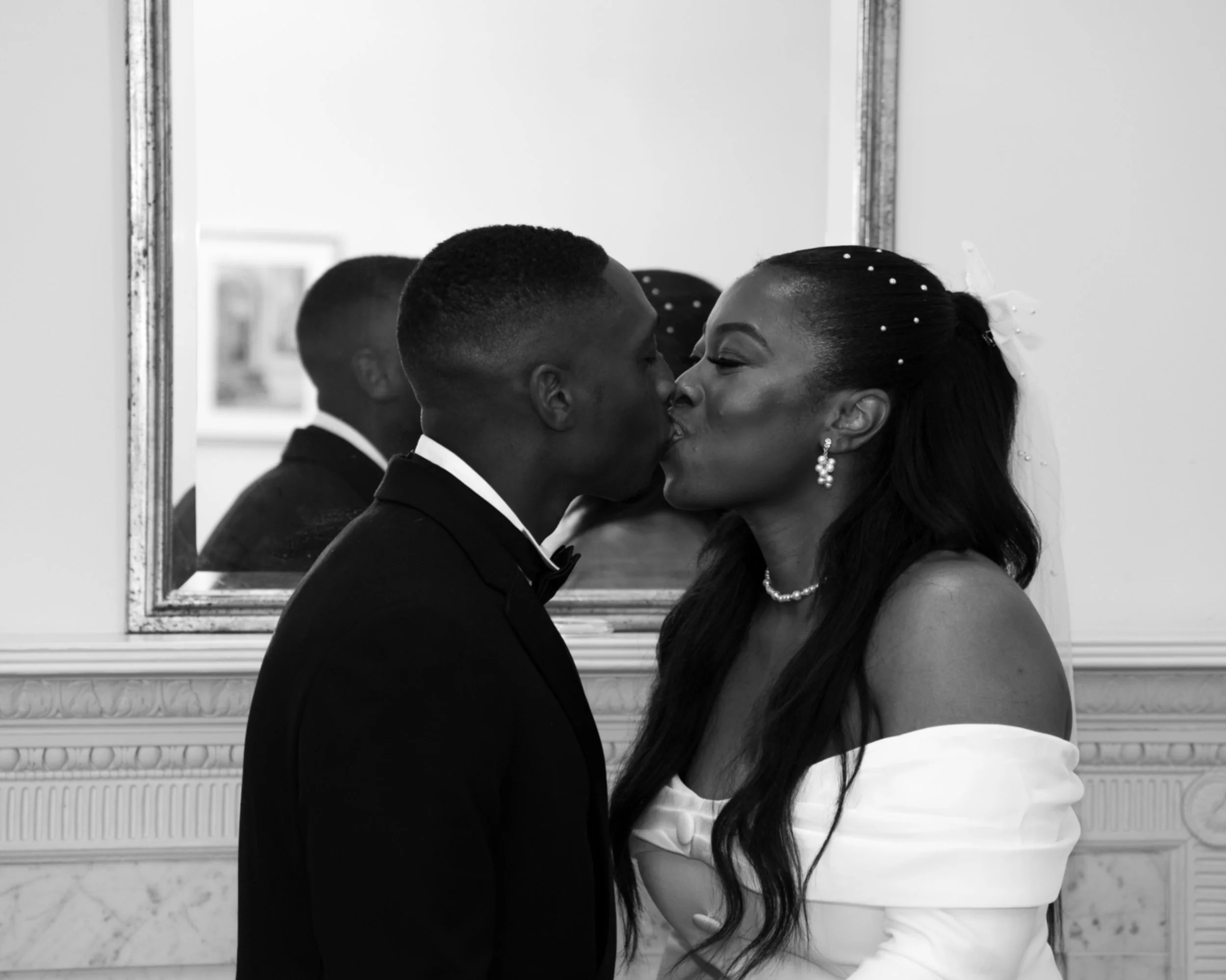 A black and white photo of a man and woman sharing a kiss in front of a mirror, dressed in formal wedding attire.