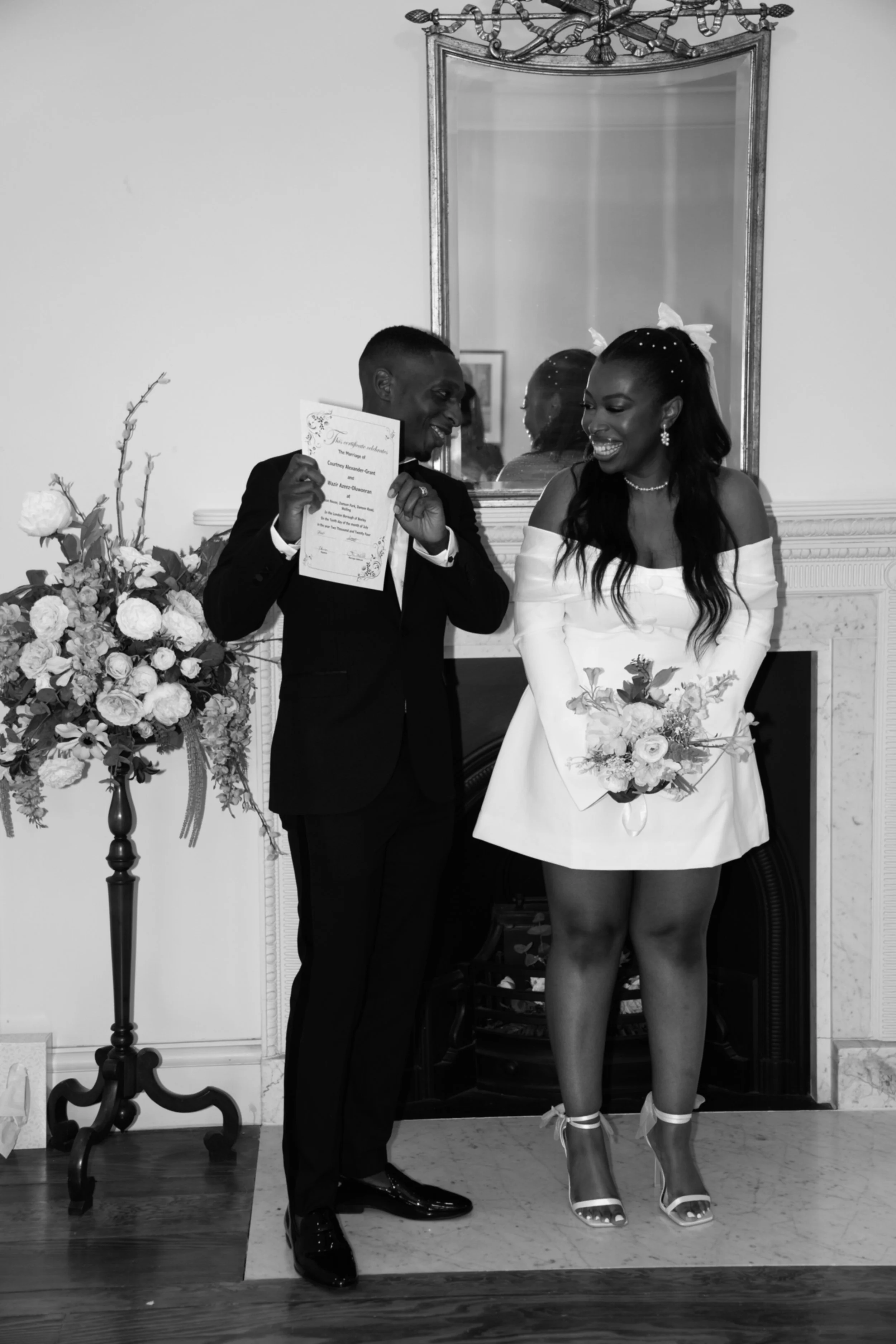 A bride in a white dress holding a bouquet and a groom in a tuxedo holding a certificate during a wedding ceremony in a decorated room.