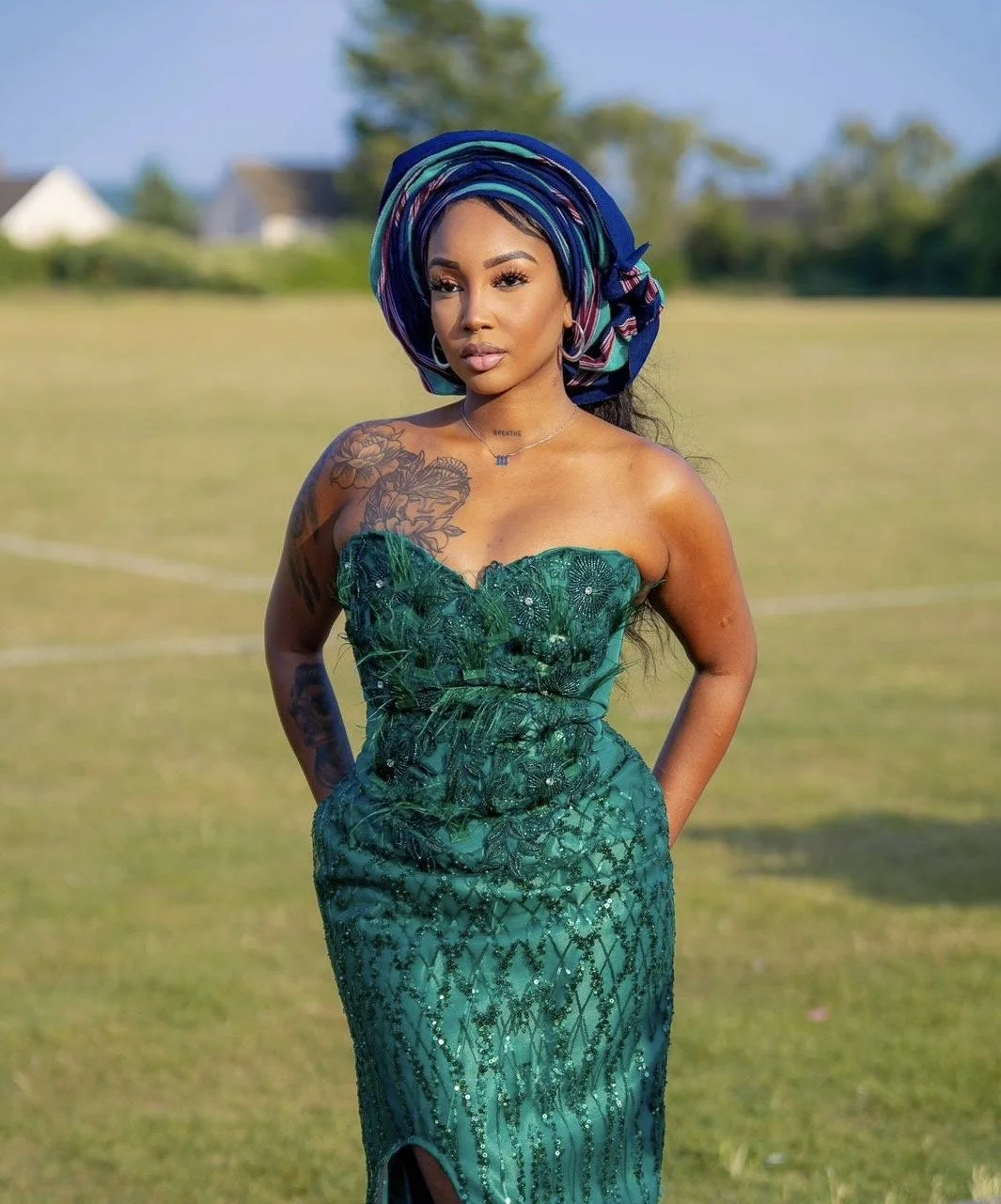 A woman standing outdoors in a grassy area with trees and houses in the background, wearing a strapless green dress with intricate patterns and a colorful headwrap.