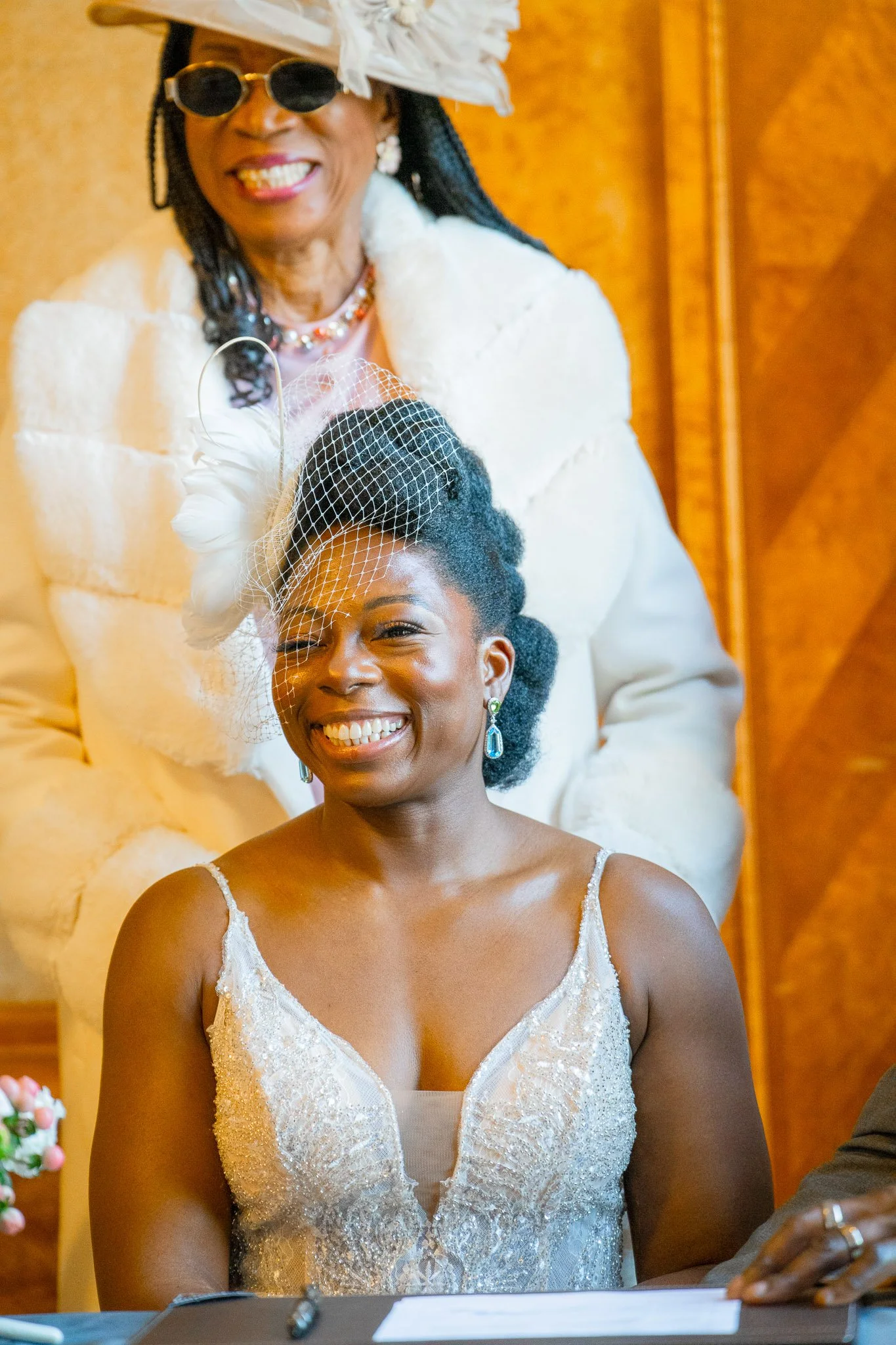 Smiling woman in wedding dress with blue earrings, seated at a table, with an older woman standing behind her, wearing a hat and sunglasses, both dressed in white for a wedding celebration.