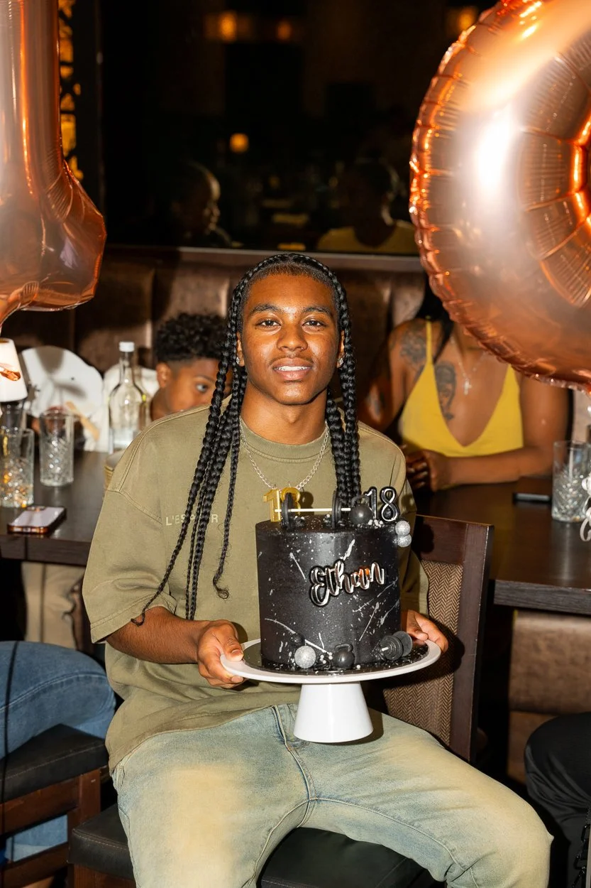 A young man with long braids holding a black birthday cake with the name 'Evan' and the year '2018' on top, celebrating his birthday in a restaurant with balloons and other guests in the background.