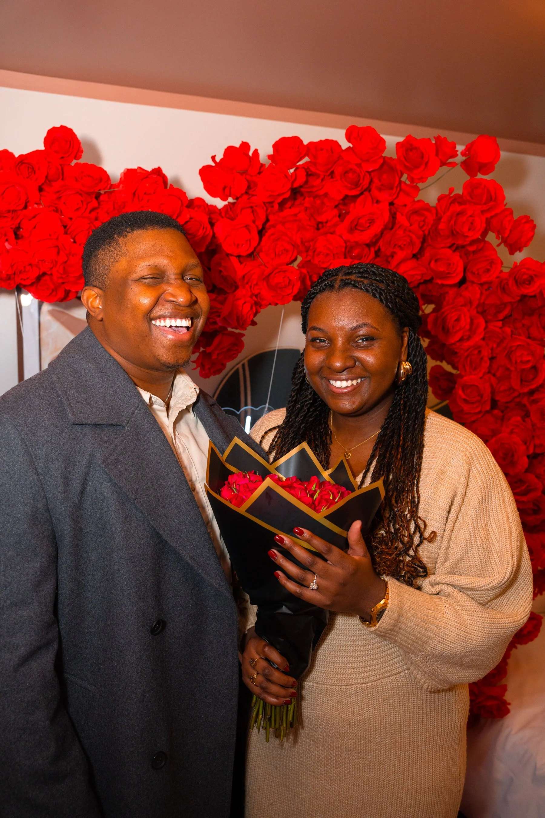 A happy couple smiling, with the woman holding a bouquet of red roses, standing against a backdrop of red roses.