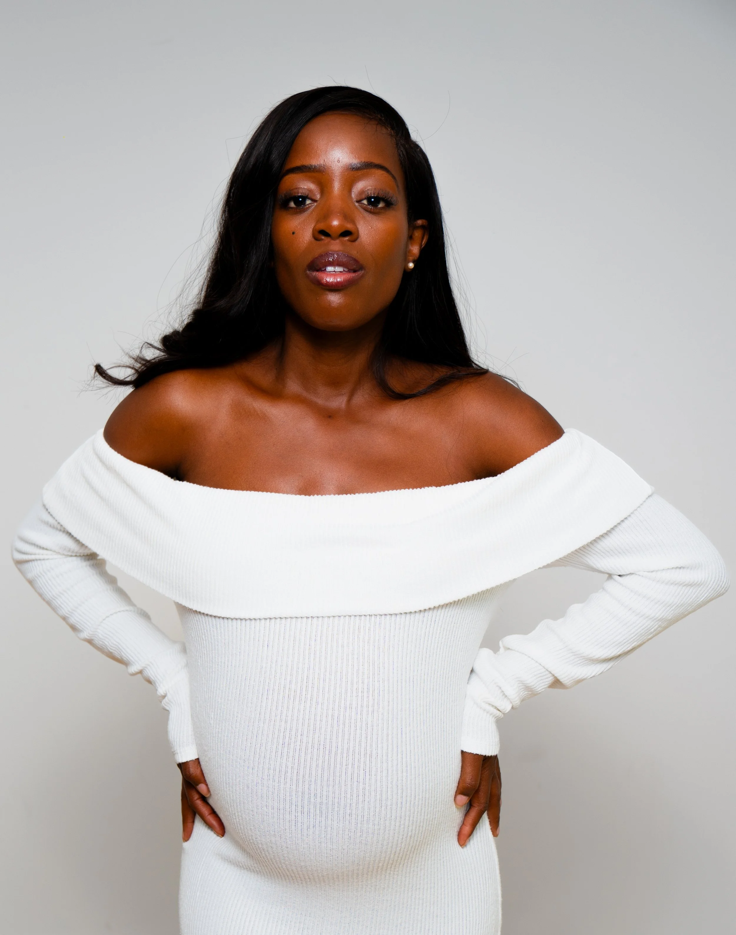 Portrait of a woman with dark hair wearing an off-shoulder white dress, standing against a plain background.