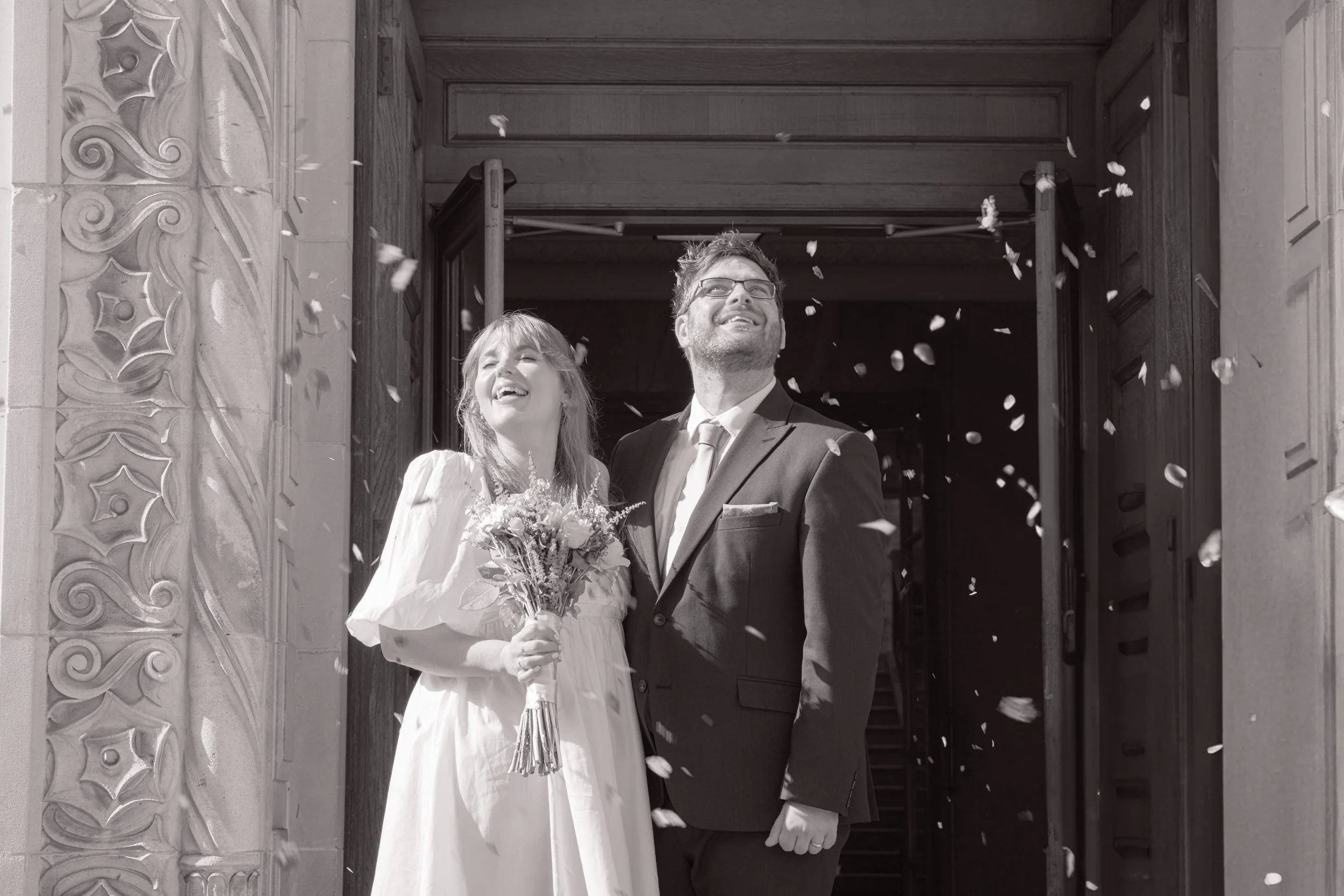 A newlywed couple standing outside a church or similar building, with petals in the air, smiling and happy, the bride holding a bouquet of flowers, black and white photo.