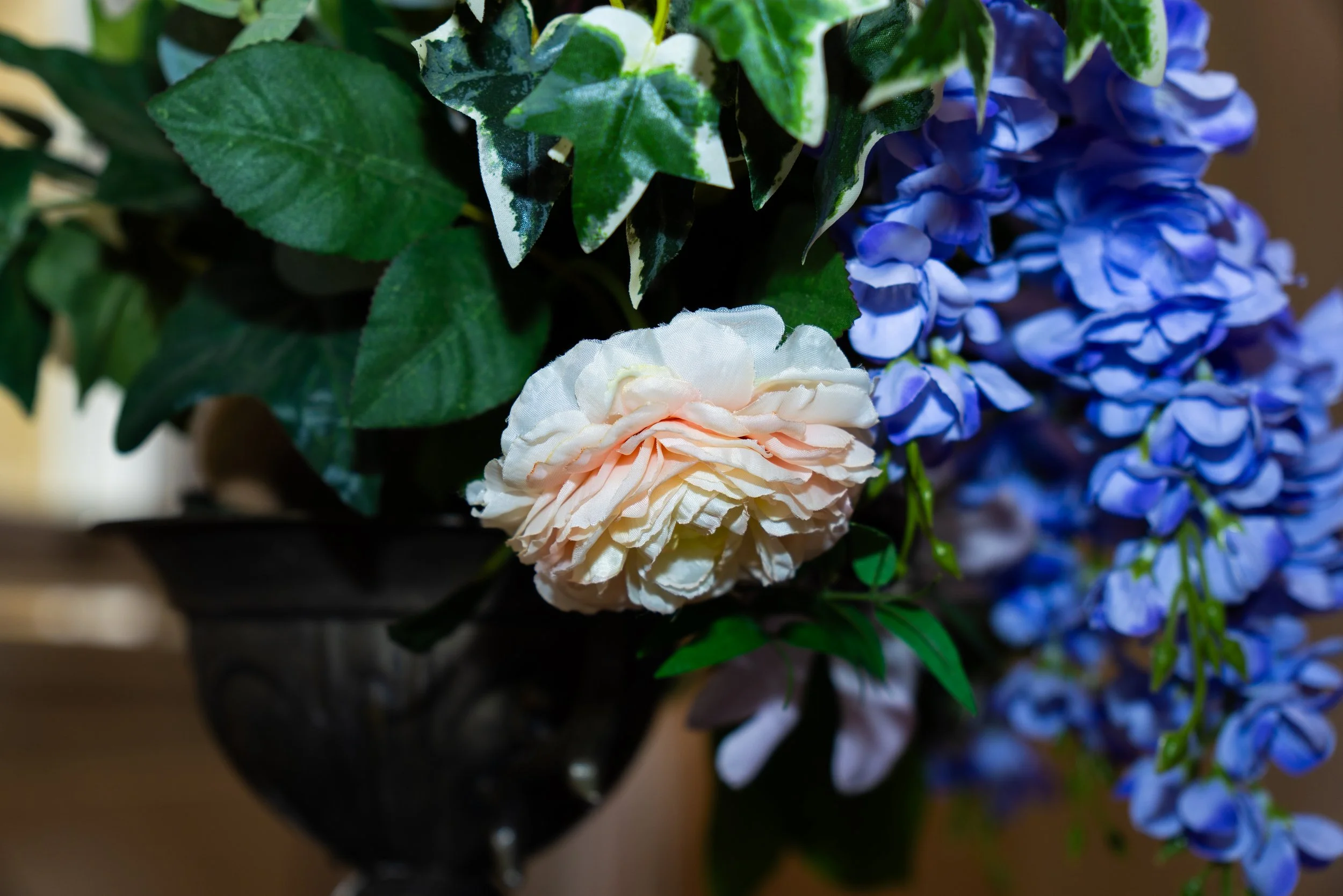 Artificial flowers in a black vase, including a peach-colored peony, blue hydrangeas, and green leaves.
