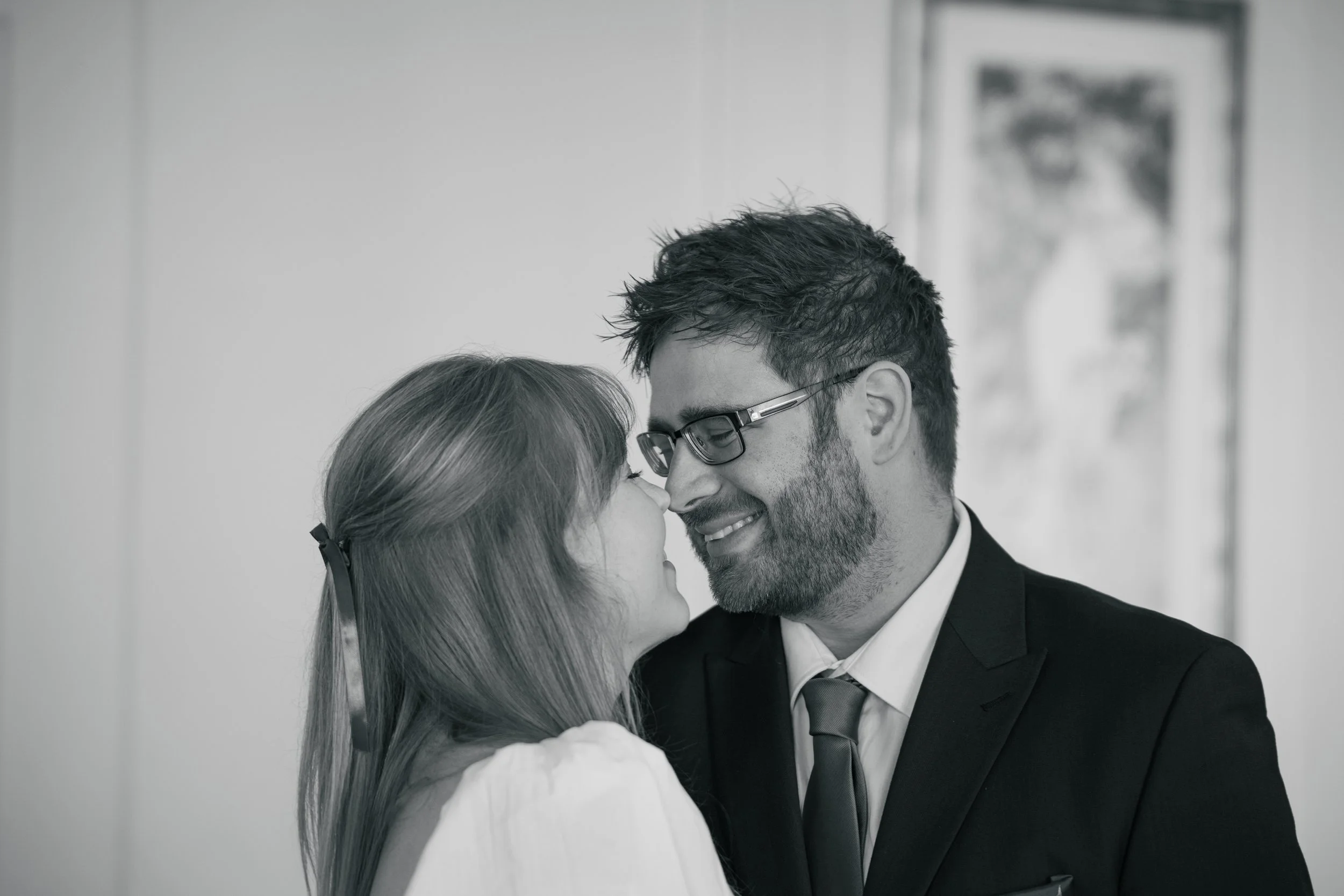 A man and woman close together, touching noses and smiling, indoors with artwork on the wall in the background.