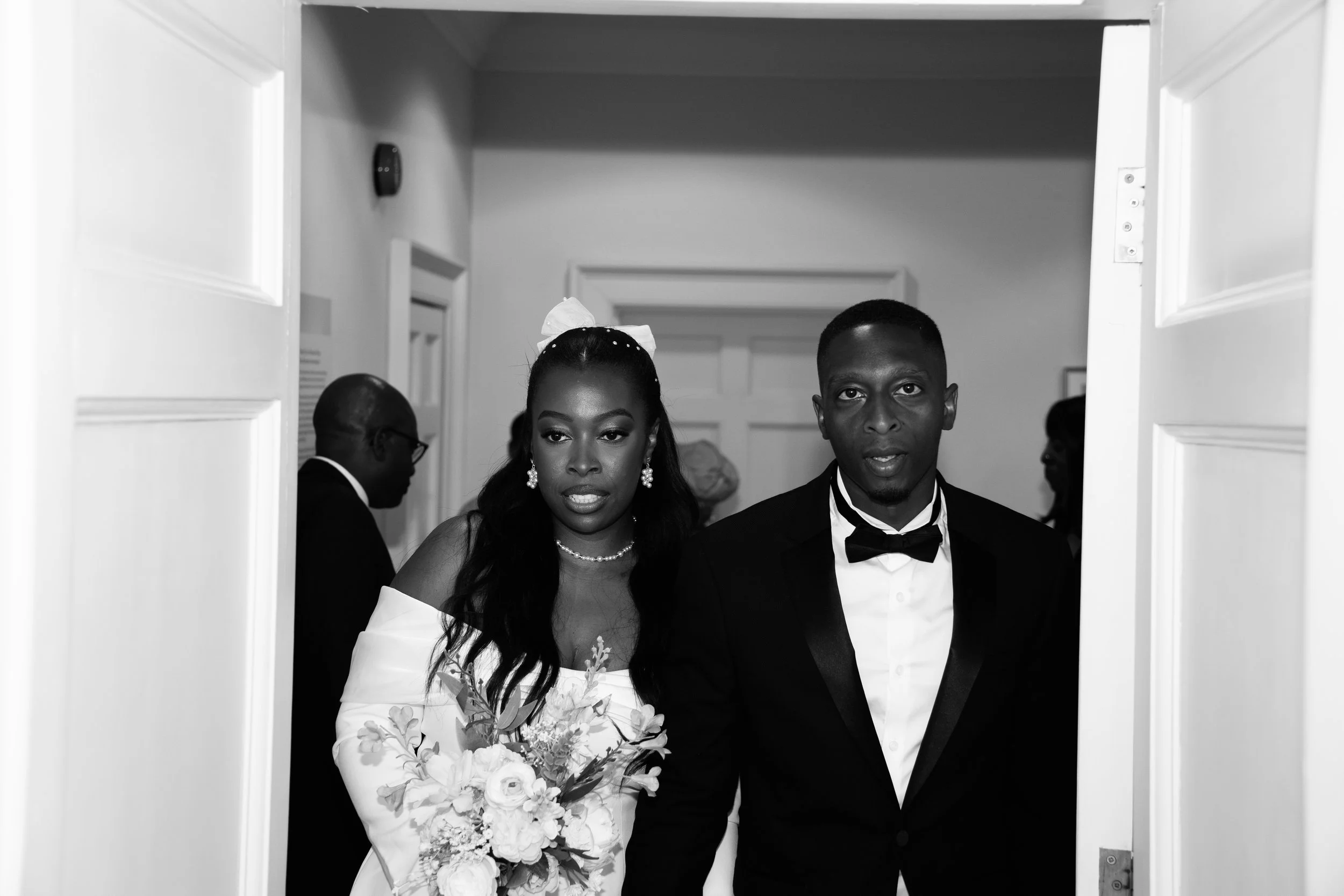 Black and white photo of a bride and groom standing in a doorway at their wedding. The bride is holding a bouquet of flowers, and the groom is wearing a tuxedo with a bow tie.