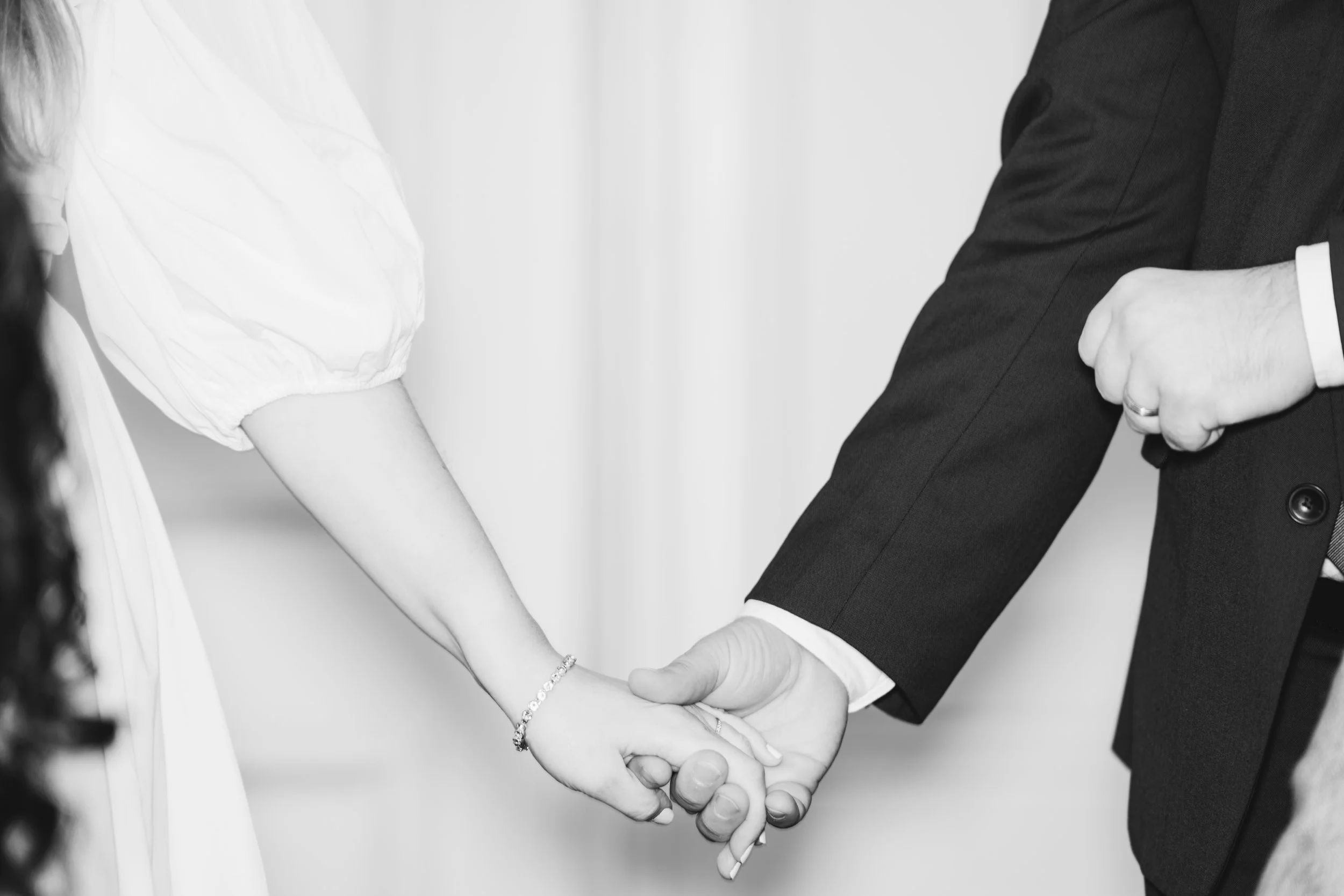 A man and woman holding hands, with the woman wearing a wedding bracelet and the man wearing a wedding band, suggesting a wedding or formal occasion.