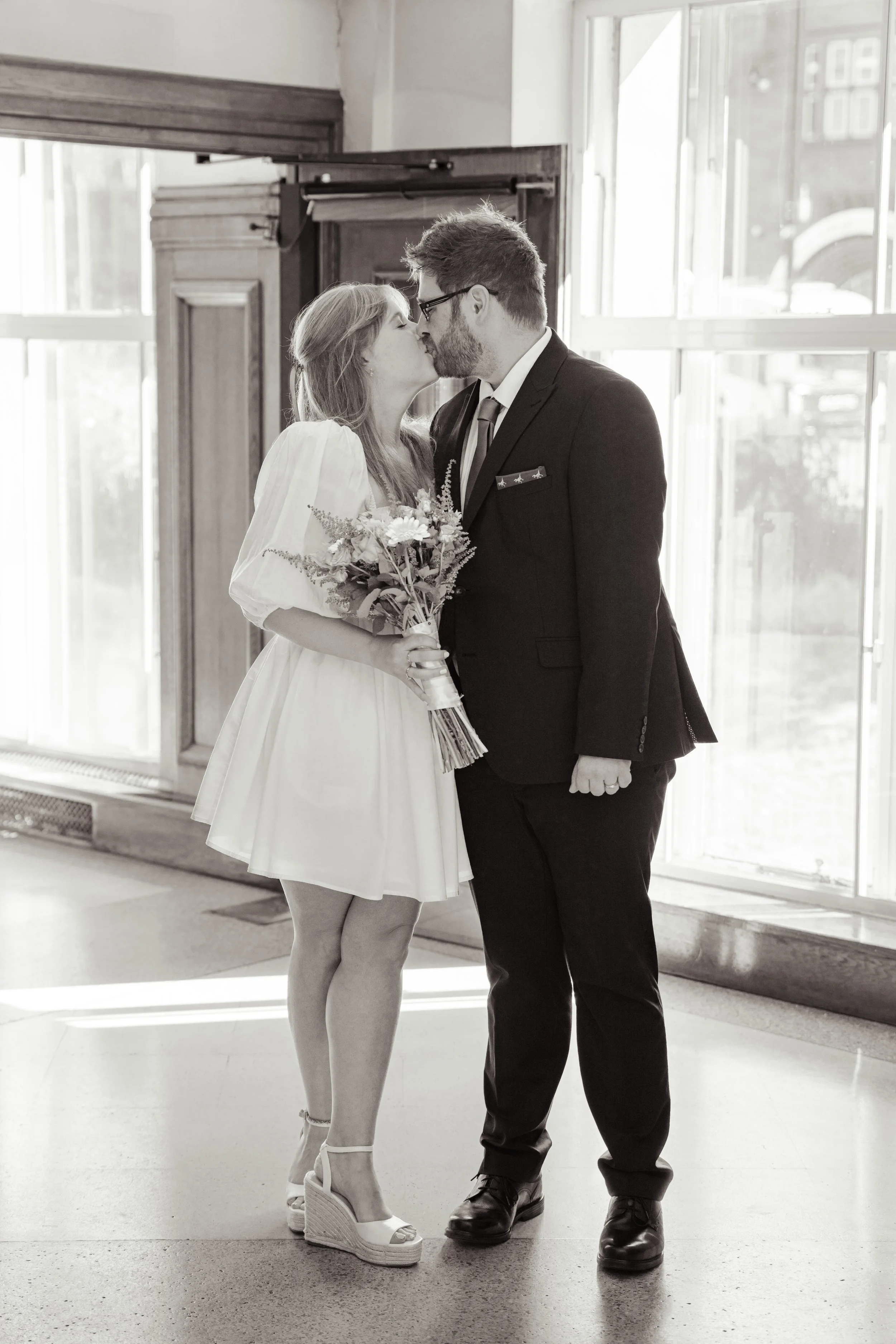 A couple in wedding attire sharing a kiss indoors near large windows, with the woman holding a bouquet of flowers.