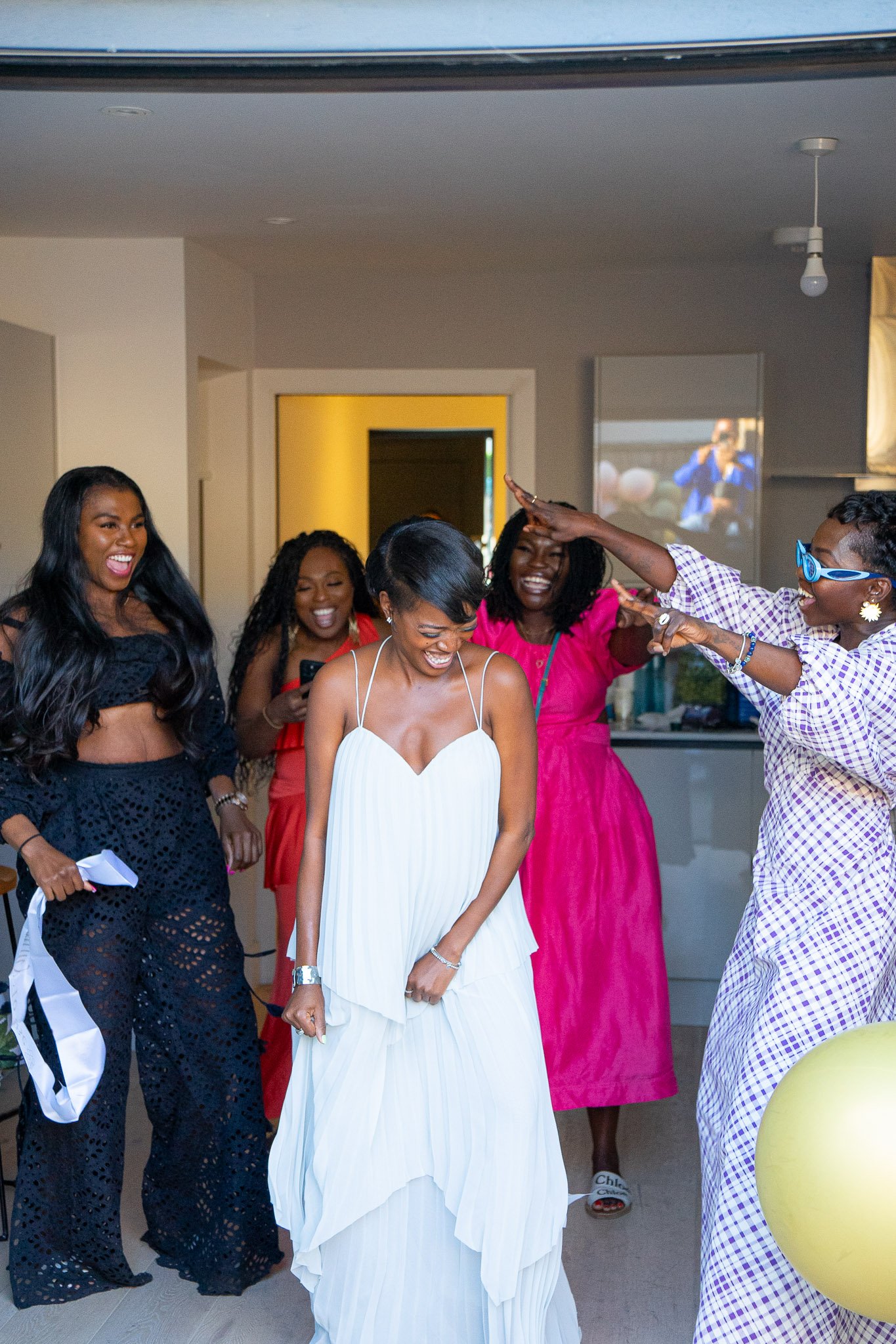A group of women dressed in colorful and elegant outfits, celebrating indoors with joyful expressions and dancing.