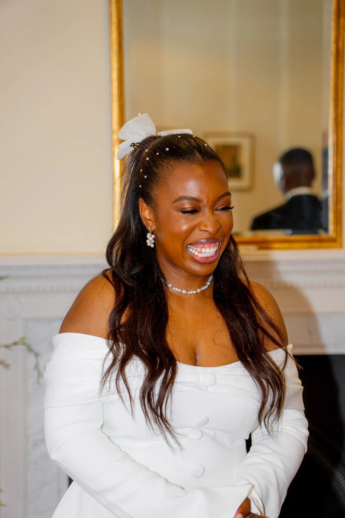 A woman in a white dress with an off-the-shoulder neckline, smiling and laughing, wearing pearl earrings and a matching pearl necklace, with a white bow in her hair, standing in a room with elegant decor and a large mirror behind her.