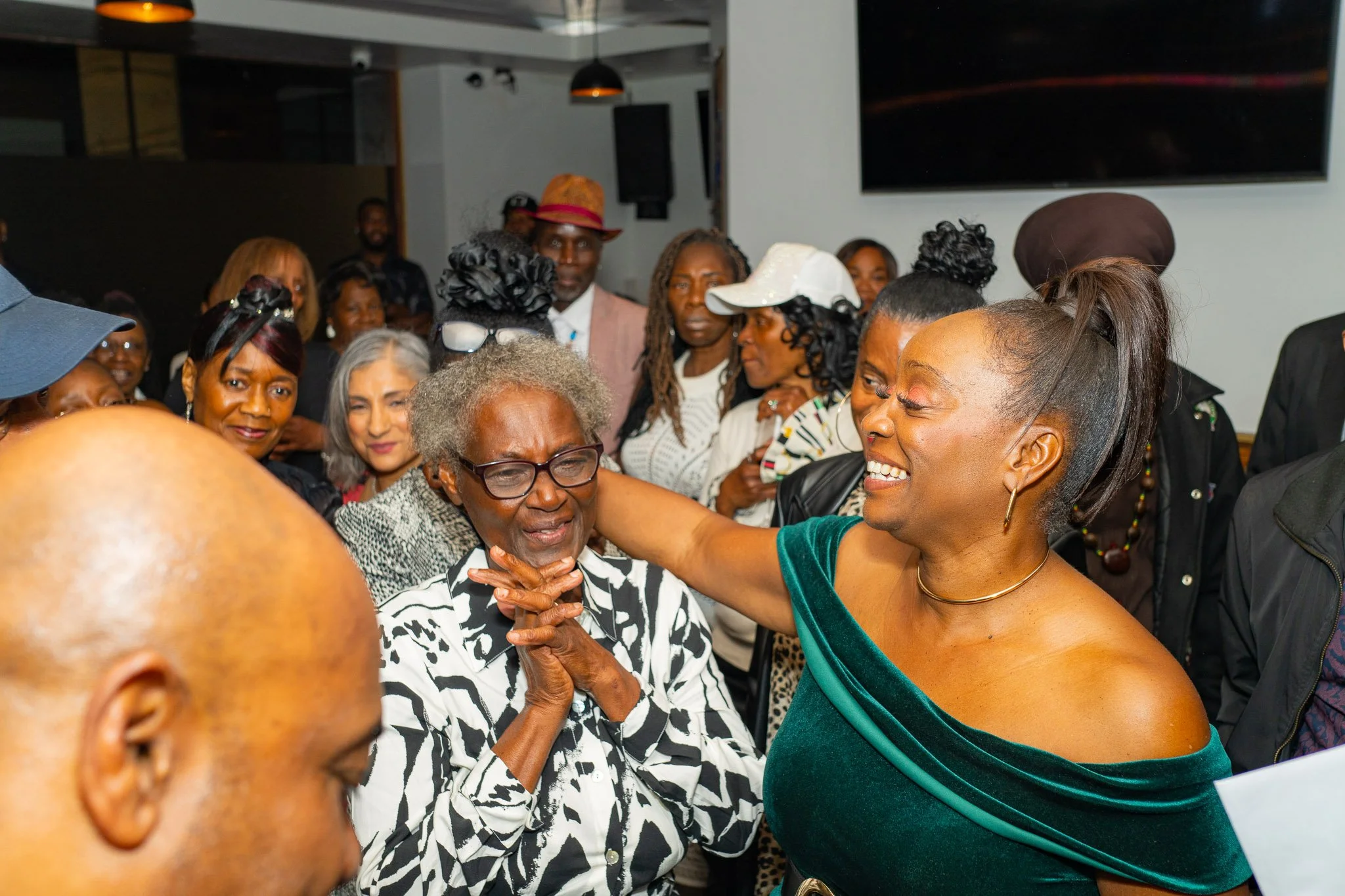A joyful group of women and men socializing indoors. An elderly woman with glasses is being embraced and smiling, while another woman with a high ponytail and green dress is smiling and talking to her. Other people are standing around, chatting and o