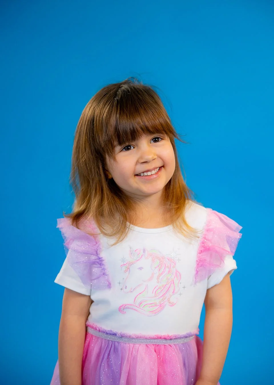 A young girl with brown hair and a big smile standing in front of a blue background, wearing a unicorn-themed dress with pink and purple accents.