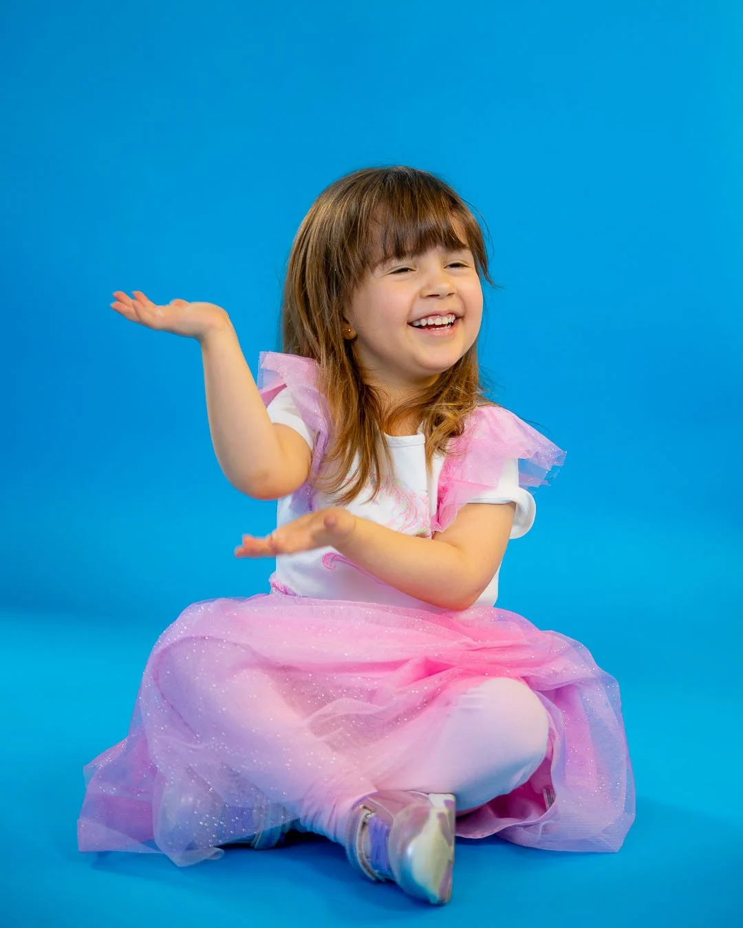 Smiling young girl dressed as a princess in a pink tutu skirt and white top, sitting on a blue background, with arms raised and a joyful expression.