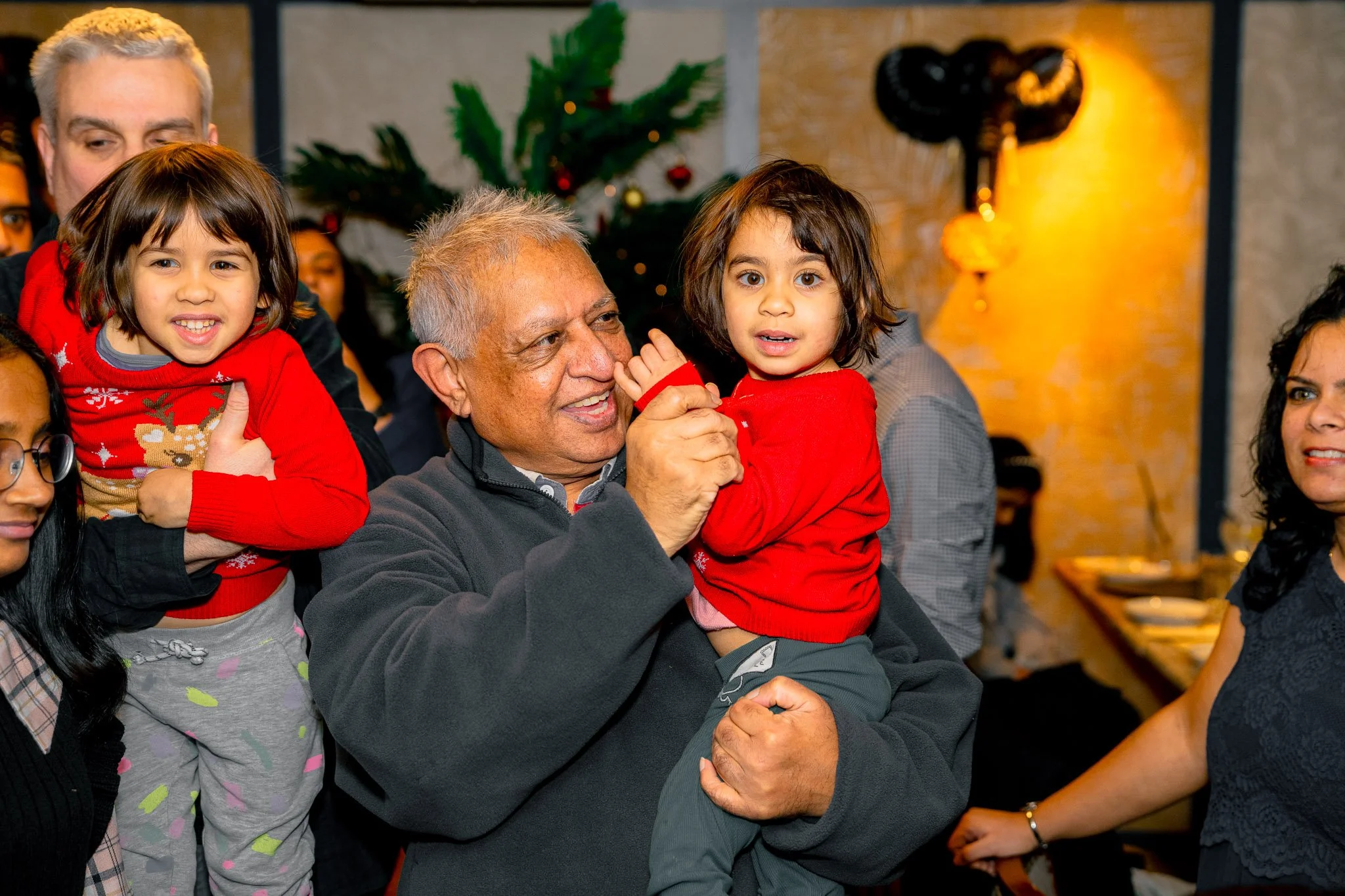 A group of people, including two young children in red sweaters, celebrating Christmas indoors with a decorated tree in the background.