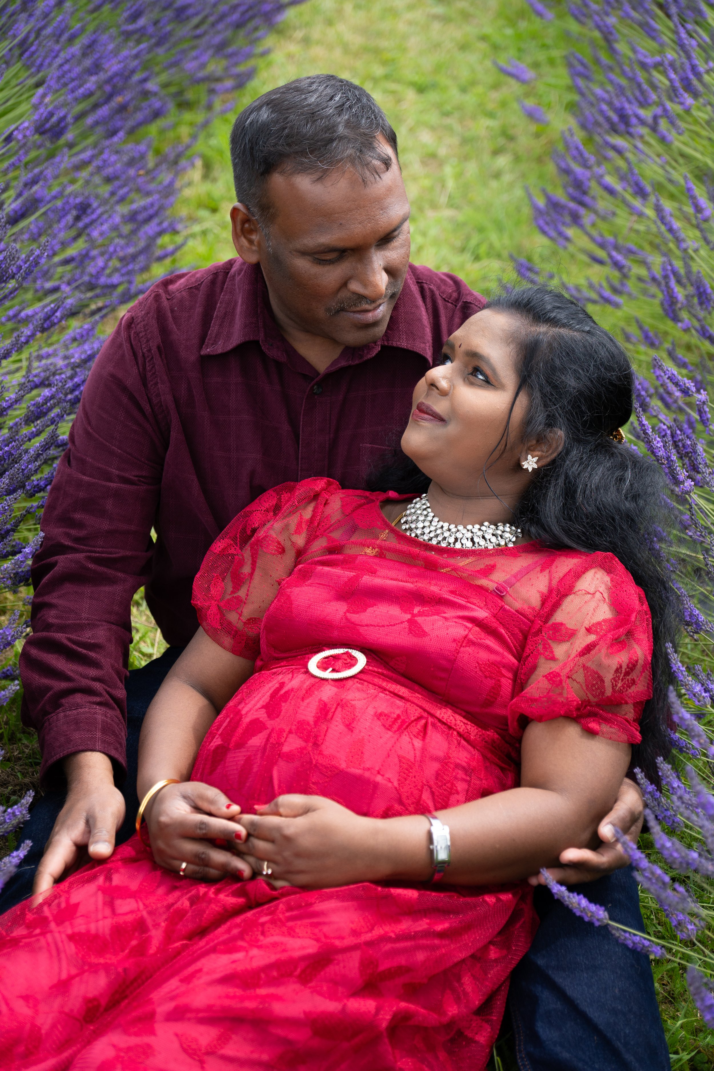 A couple sitting in a lavender field, with the woman pregnant and wearing a red dress, jewelry, and a wristwatch, while the man looks at her lovingly.