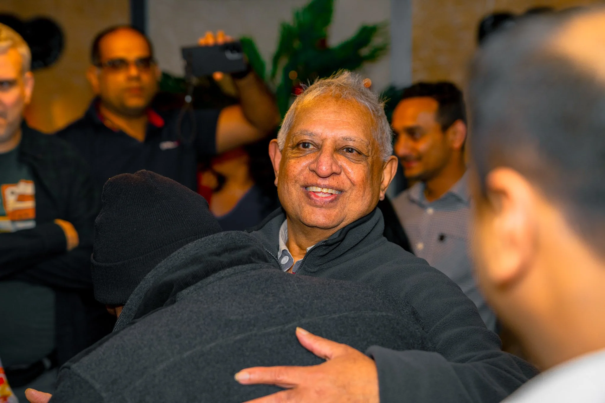 A smiling man with gray hair embraces another person in a black hoodie during a social gathering, with several people in the background, some talking and one person taking a photo.