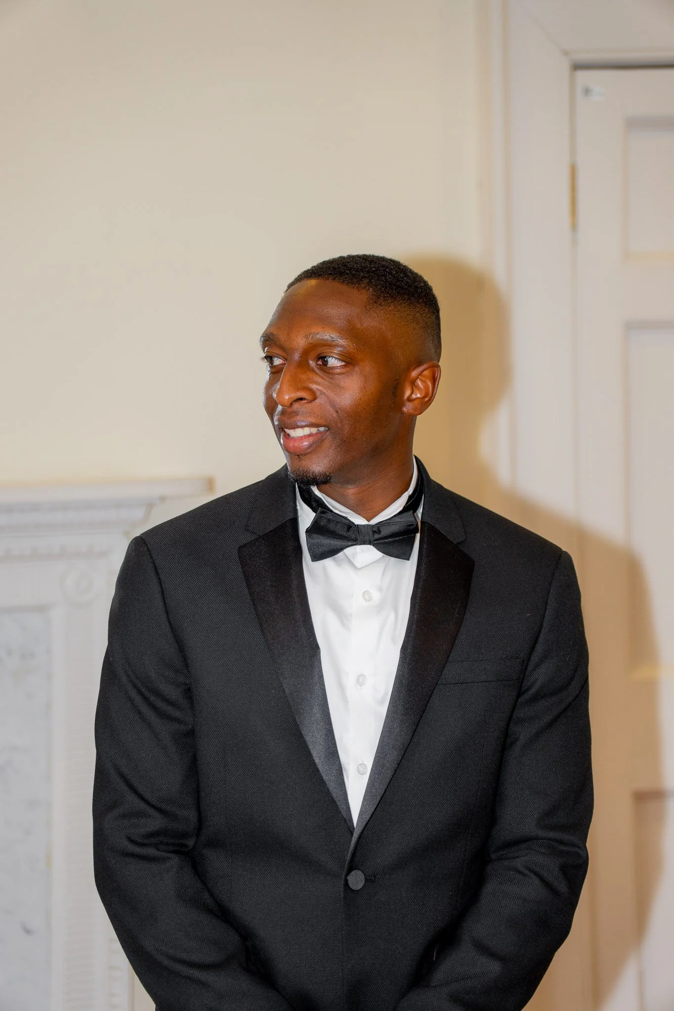 A man dressed in a black tuxedo with a white dress shirt and black bow tie, standing indoors near a white fireplace and door, looking to the side.