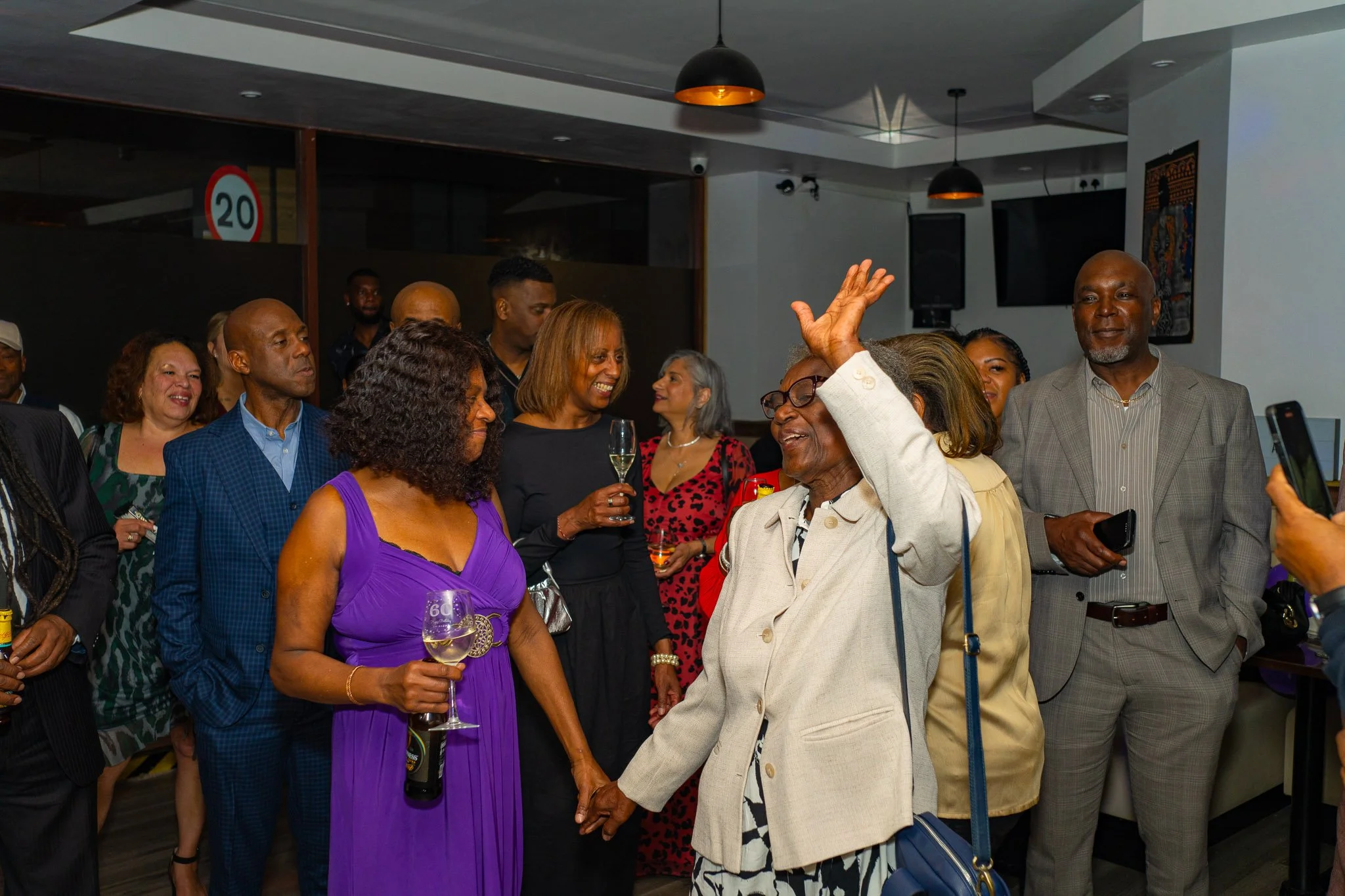 A group of diverse people at a celebration or social gathering, some holding drinks, engaging in conversation, and smiling, in an indoor setting.