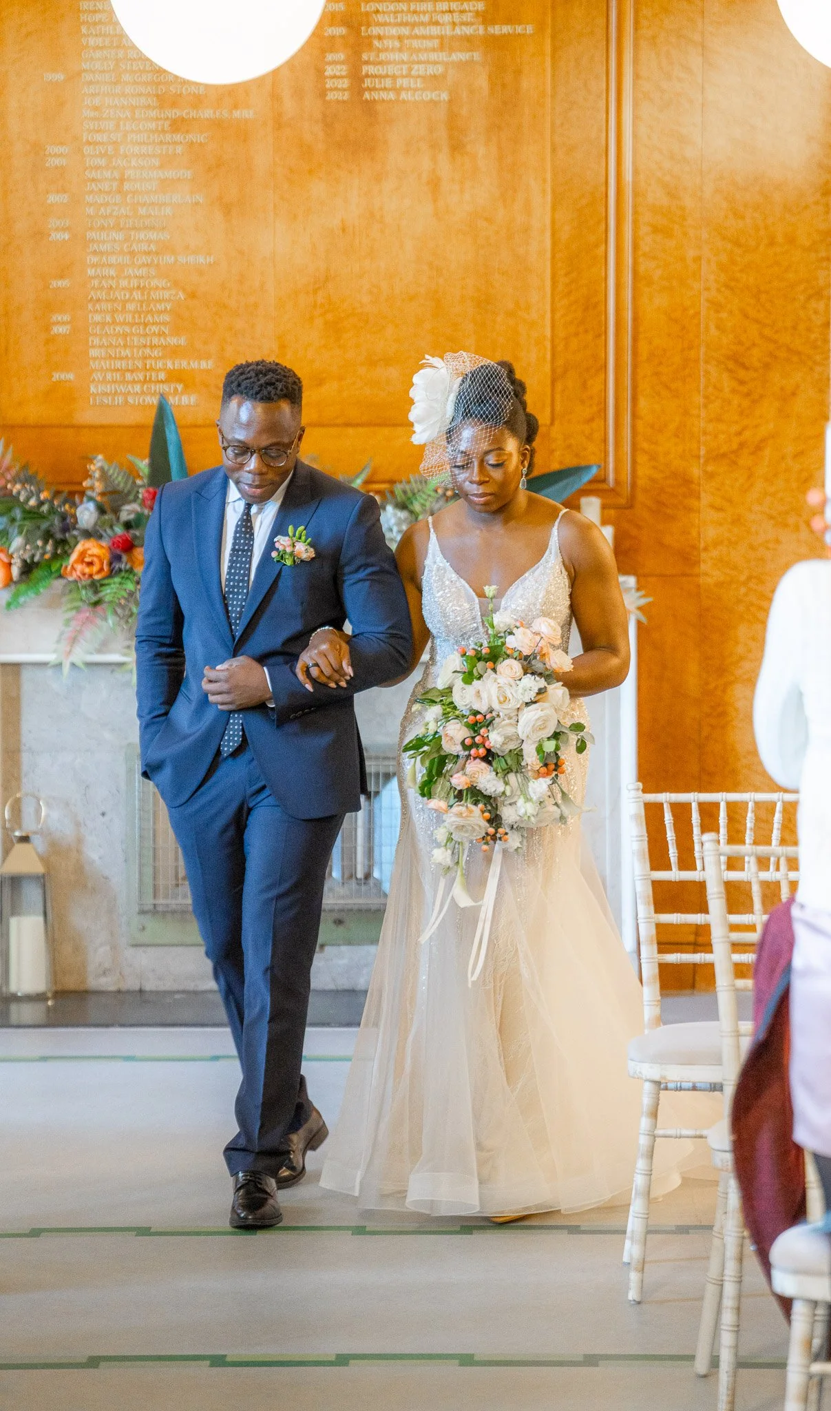 A bride in an elegant white wedding gown holding a bouquet of white and peach roses, walking arm-in-arm with a man in a dark suit with glasses, at a wedding ceremony in an indoor venue with wooden walls and floral decorations in the background.