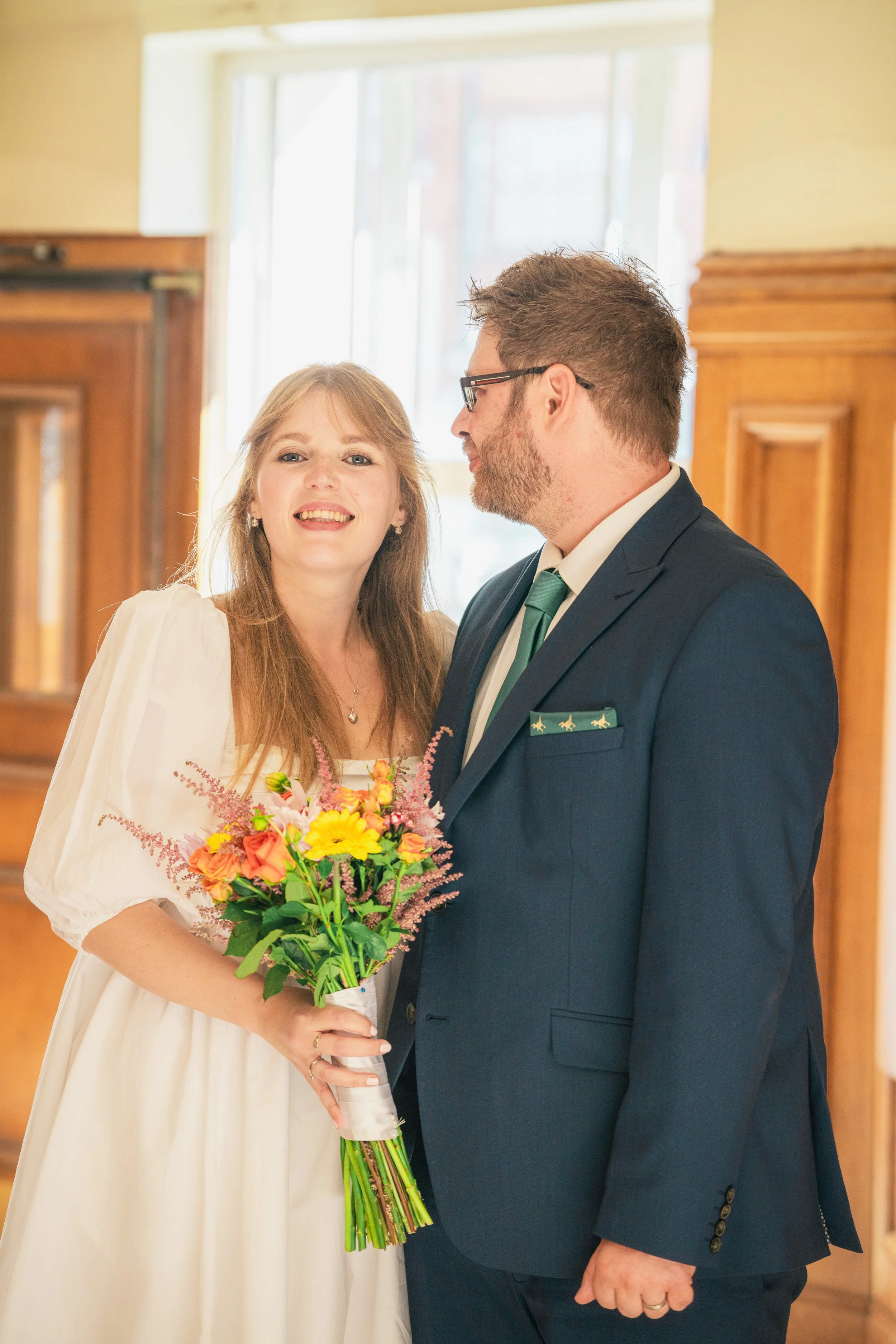A newlywed couple stands together indoors, the woman holding a colorful bouquet, both smiling and dressed in wedding attire.