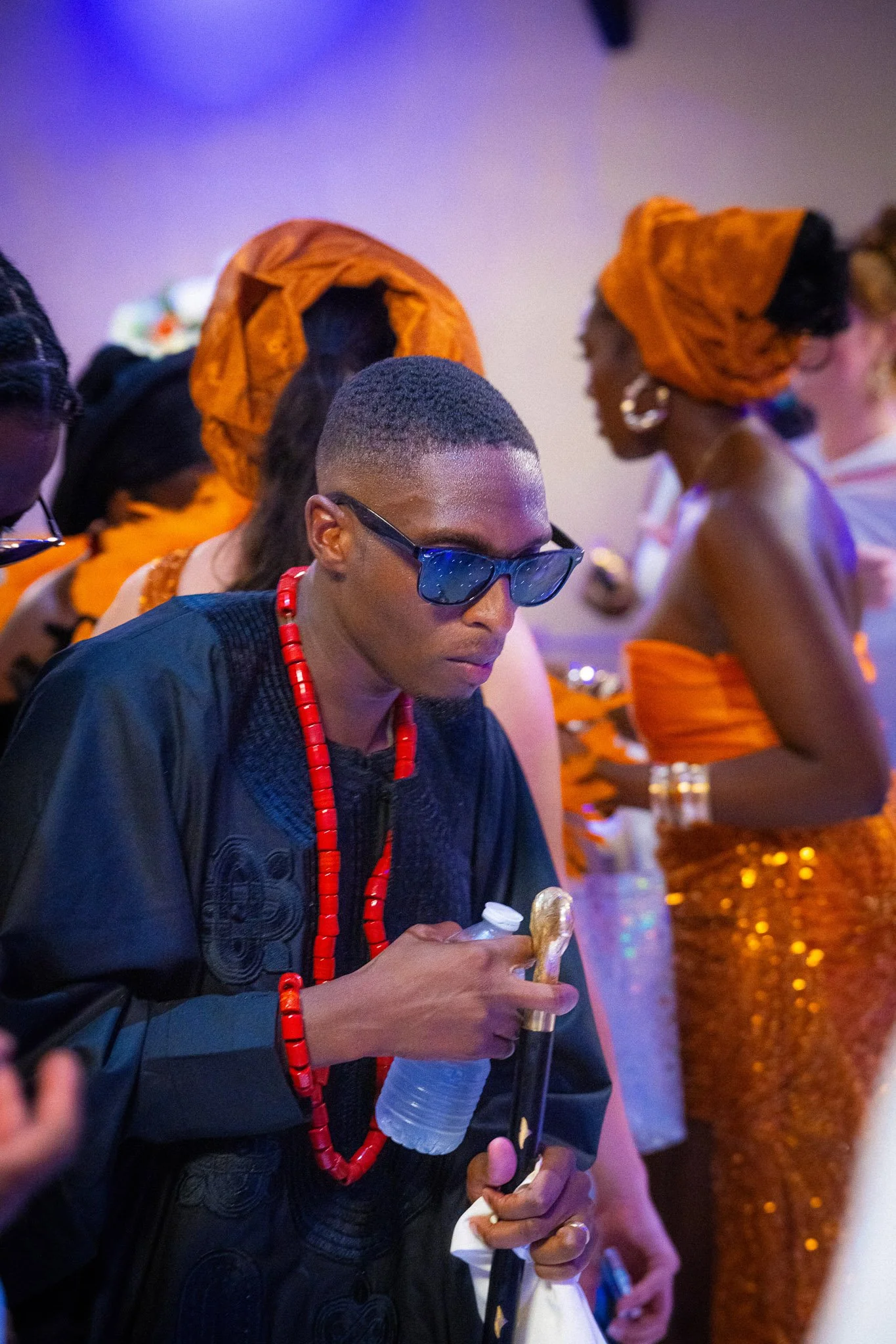 A woman with short hair wearing sunglasses, traditional jewelry, and African attire holding a water bottle and walking with other women dressed in orange headwraps and dresses at an event.